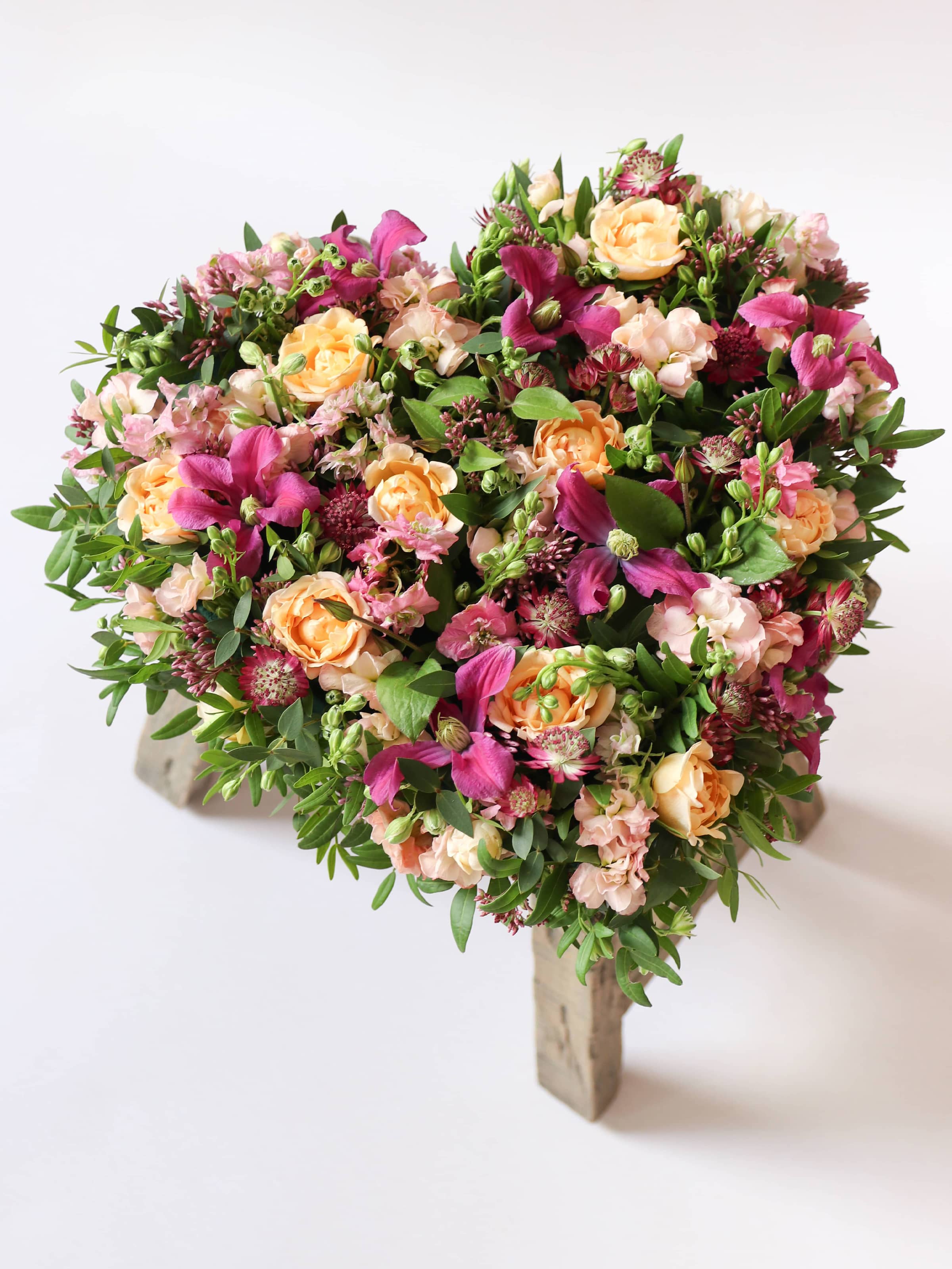 Heart-shaped funeral posy with bright pink and peach flowers on a white background sitting on top of a wooden stool.