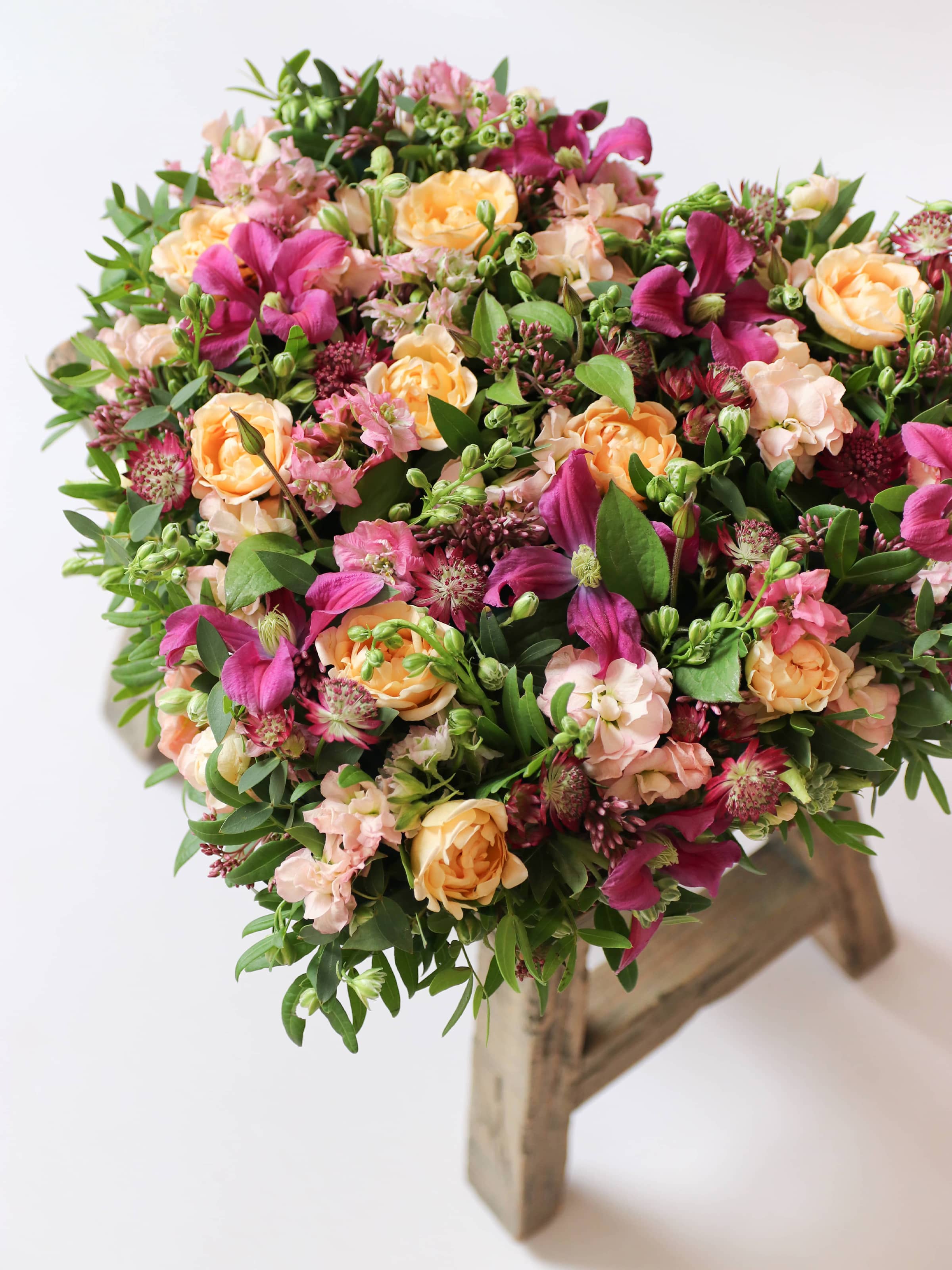 Heart-shaped posy with bright pink and peach flowers on a white background, sitting atop a wooden stool.