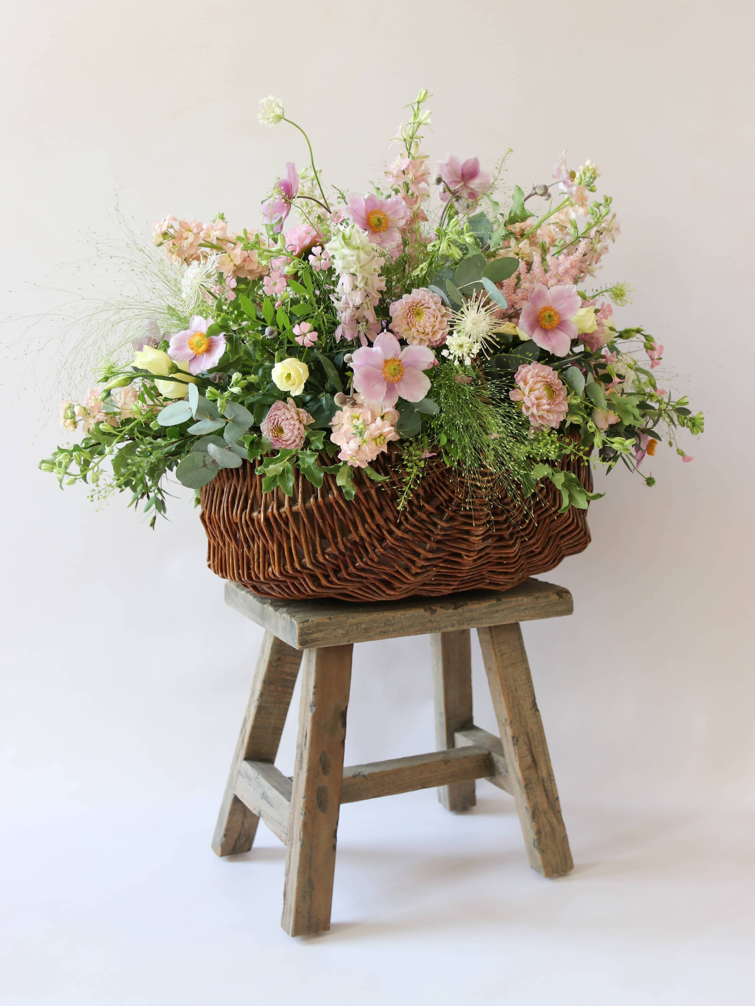 A side view of a wicker basket arrangement featuring pastel pink and yellow flowers. The basket arrangement is placed on a rustic wooden stool against a neutral background and features pink Dahlias, yellow Lisianthus and peach Stocks.