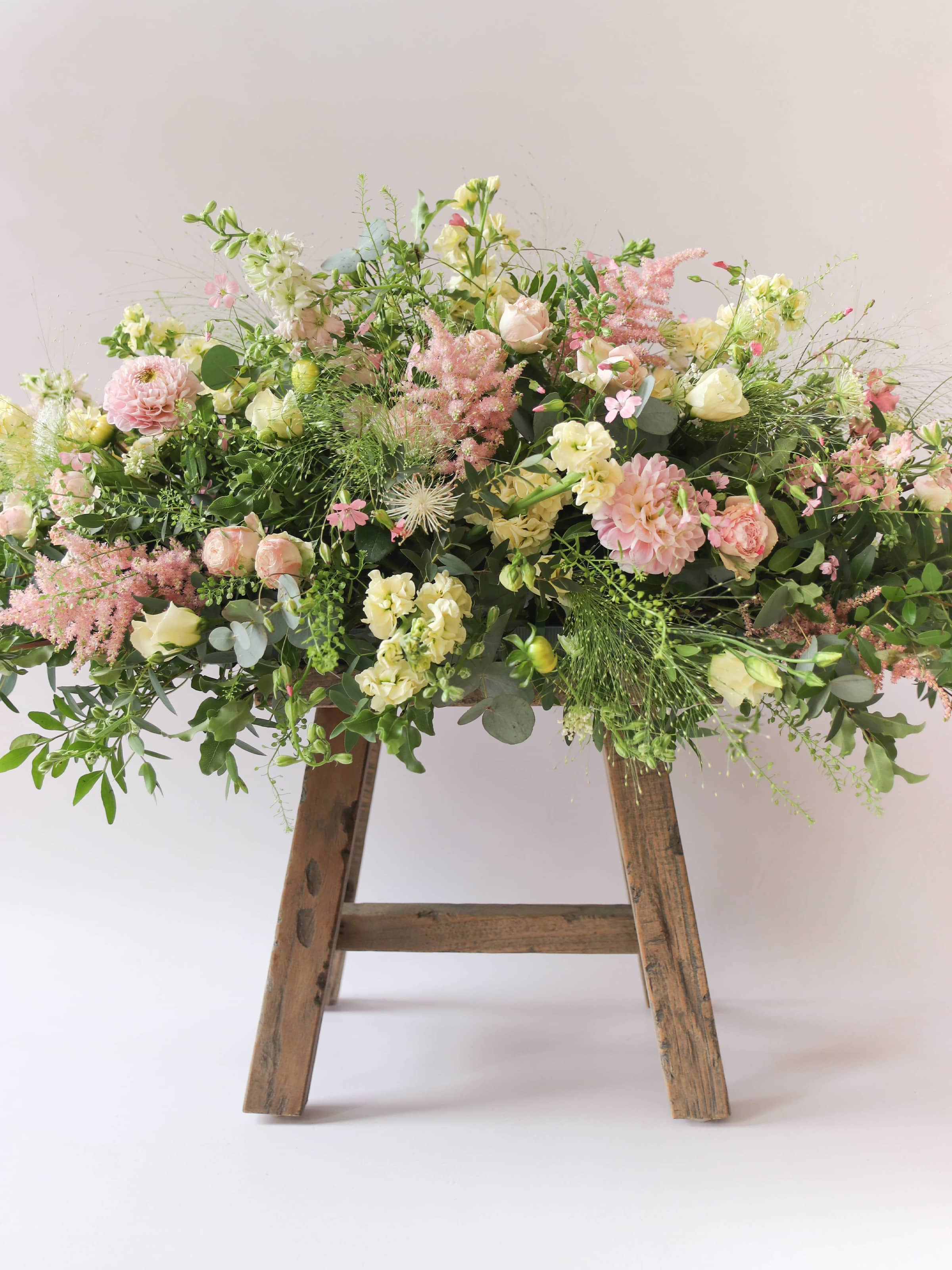 A side view of a 3ft coffin spray featuring pastel pink, yellow and peach flowers and foliage. The funeral arrangement is sitting on a rustic wooden stool against a neutral background and features Roses, Astlibe, Stocks and Lisianthus.