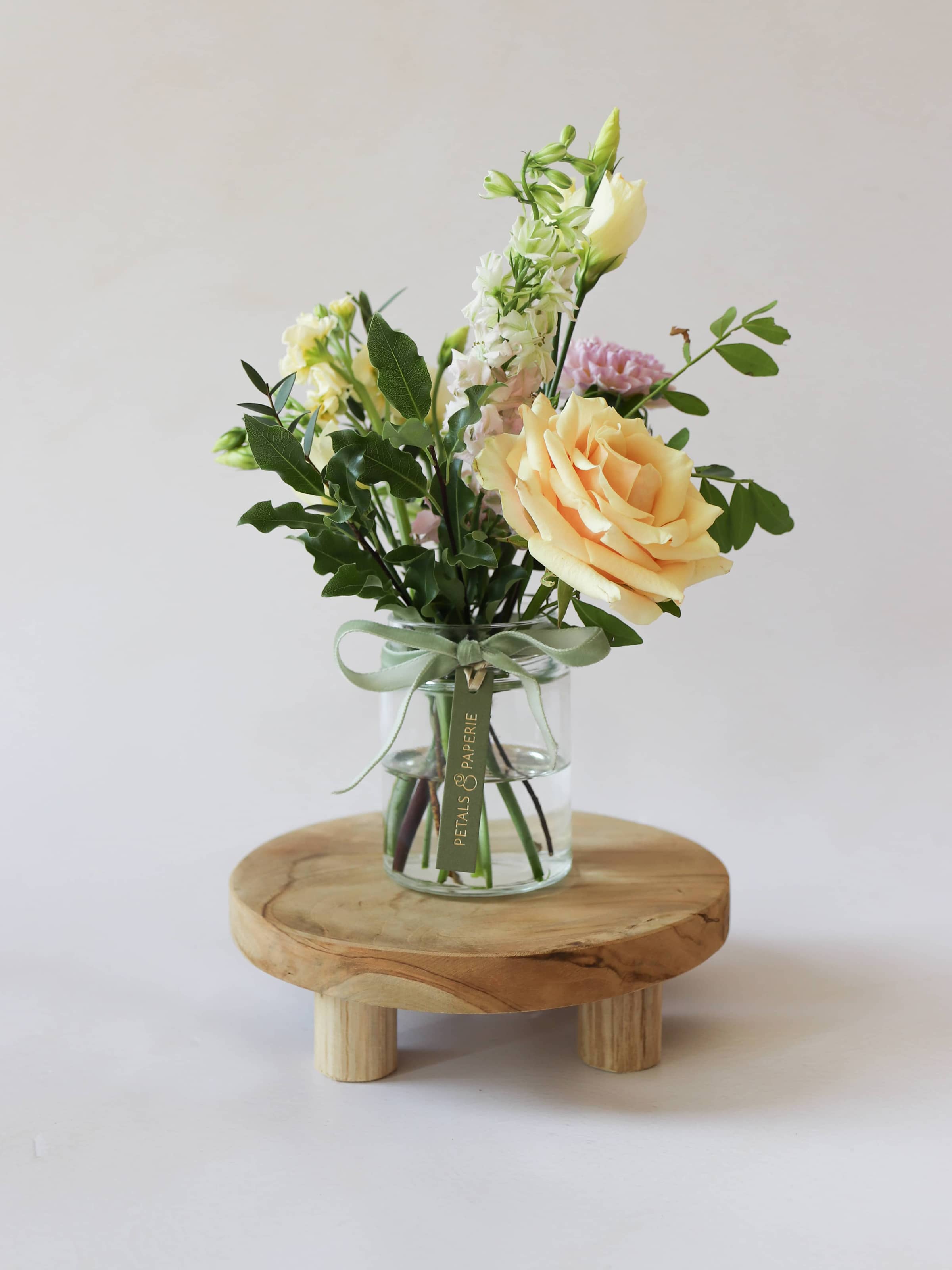 A single jam jar with pastel coloured flowers sitting on a wooden stool against a light background