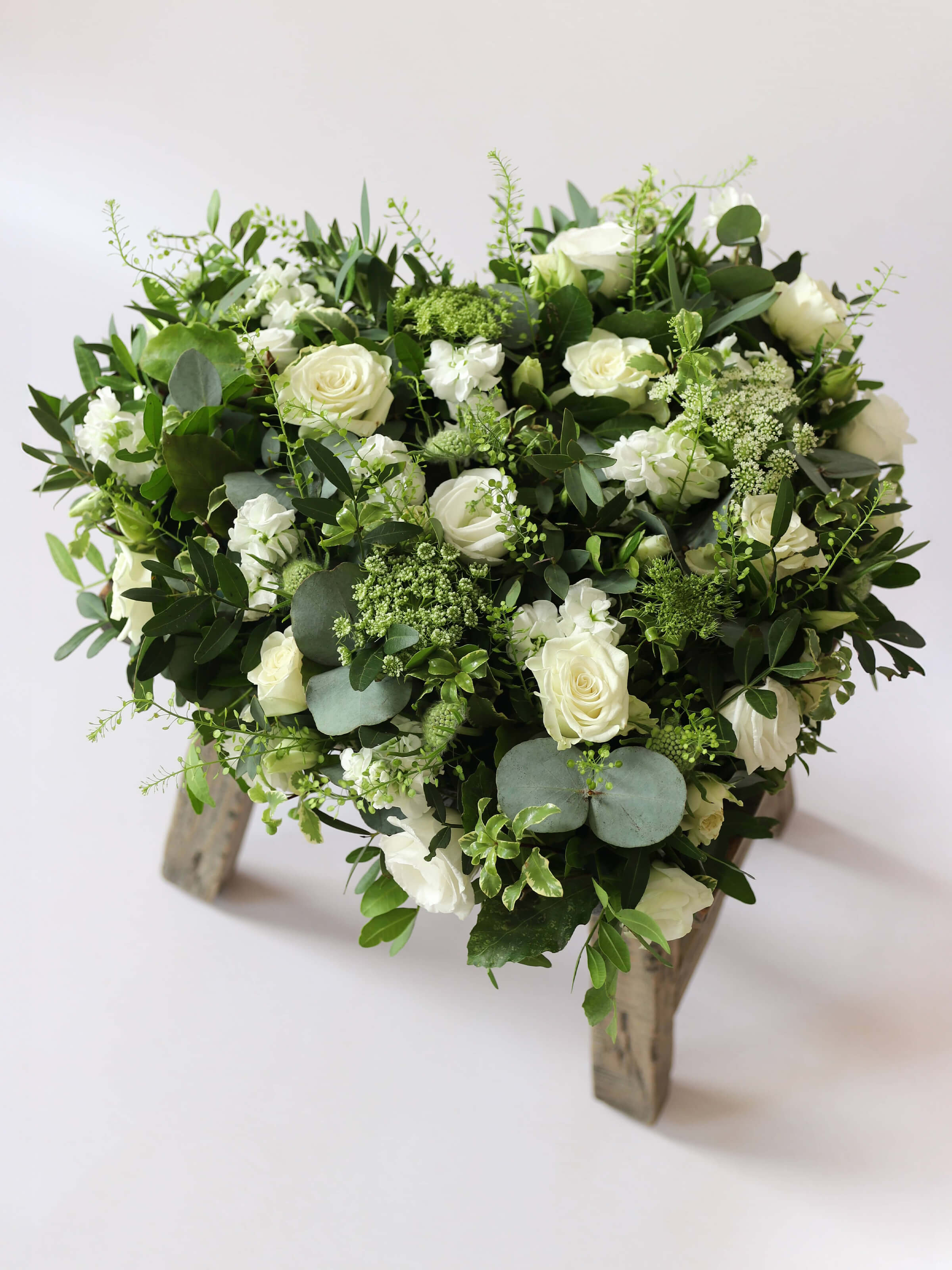 Heart shaped funeral posy with white and green flowers on a white background sitting on a wooden stool