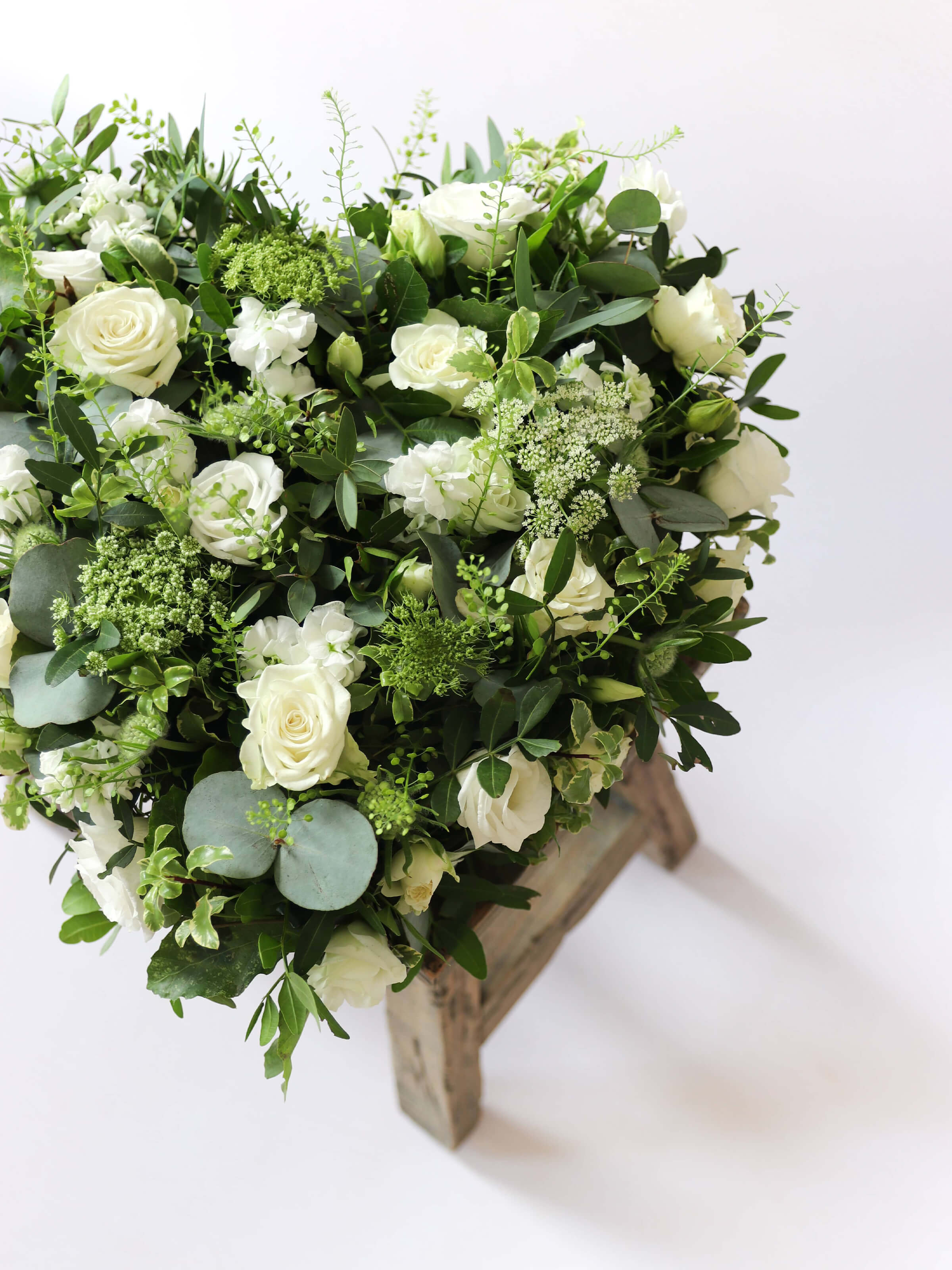 Close up of a Heart shaped funeral posy with white and green flowers on a white background sitting on a wooden stool