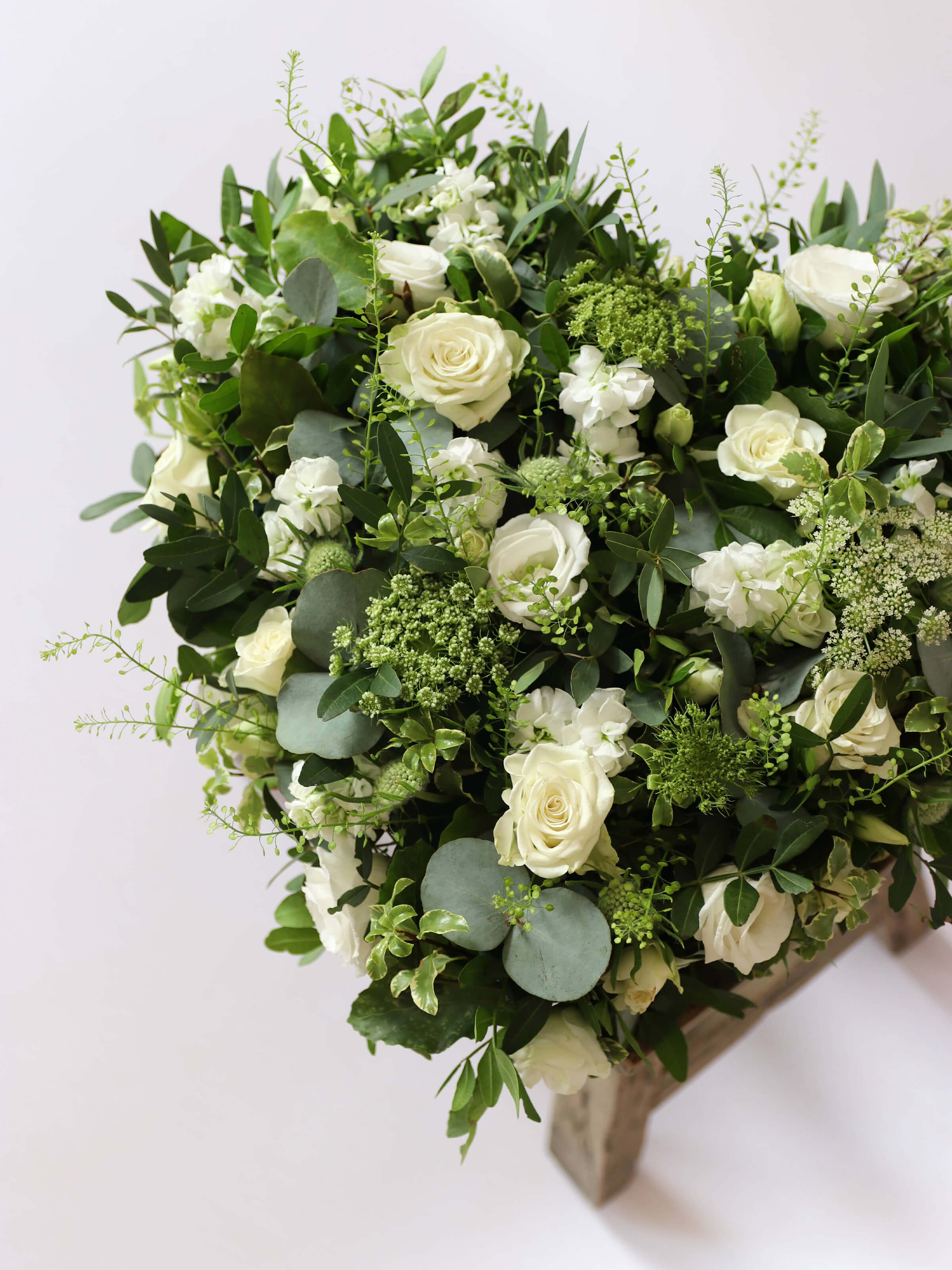 Heart shaped funeral posy with white and green flowers on a white background sitting on a wooden stool
