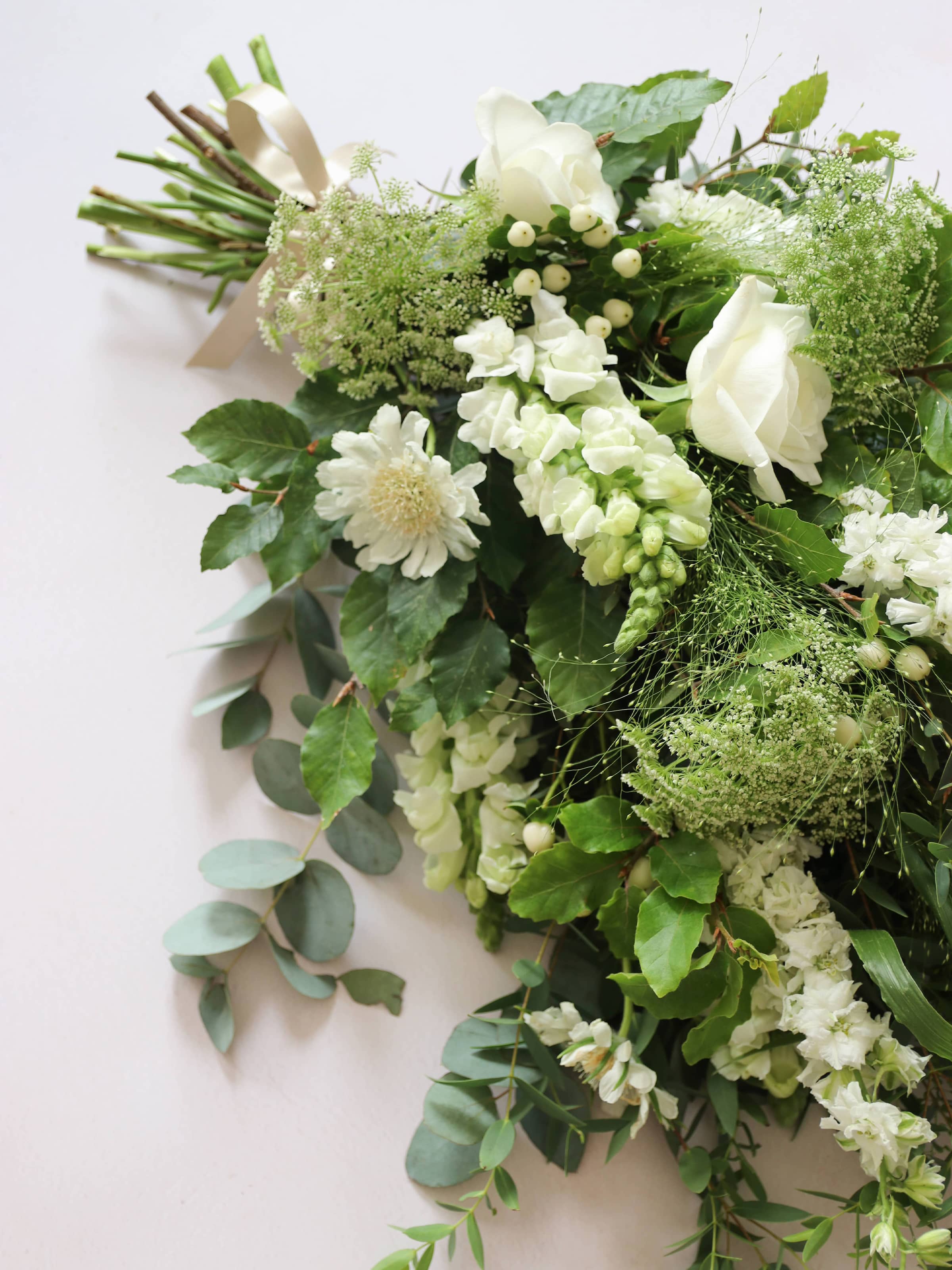 A close up view of a white and green tied funeral sheaf containing white flowers and green foliages, tied with a champagne colour satin ribbon. The sheaf contains Delphiniums, Snapdragons, Ammi and Roses.