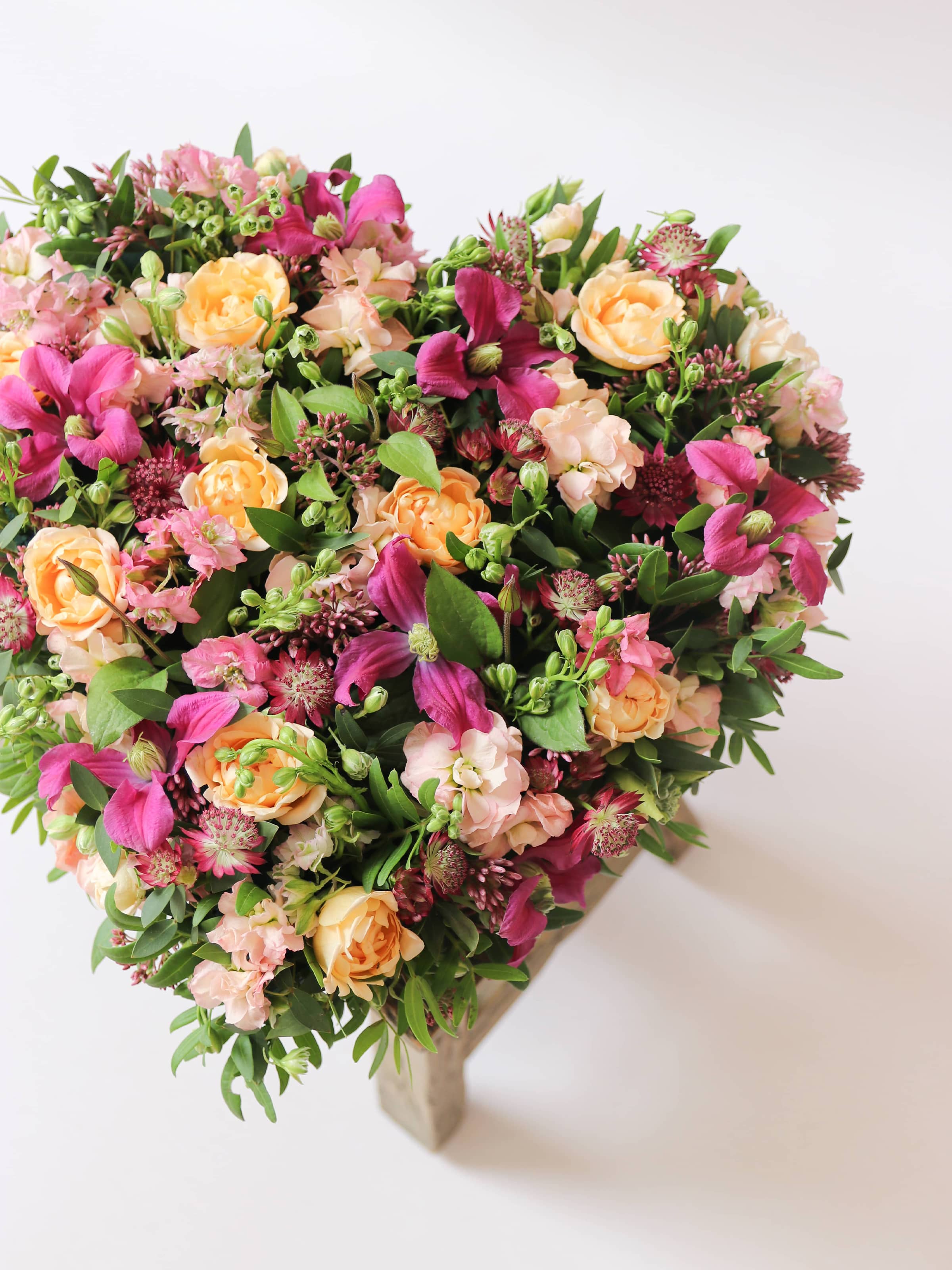 Top down, off canvas heart-shaped funeral posy with bright pink and peach flowers on a white background on top of a wooden stool.