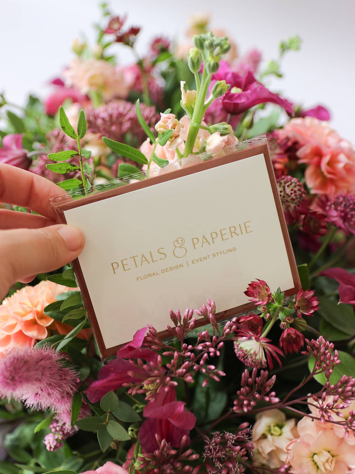 Person holding a message card with 'Petals & Paperie' branding in a bouquet of flowers