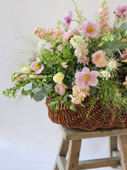 A side view of a wicker basket arrangement featuring pastel pink and yellow flowers. The basket arrangement is placed on a rustic wooden stool against a neutral background and features pink Dahlias, yellow Lisianthus and peach Stocks.
