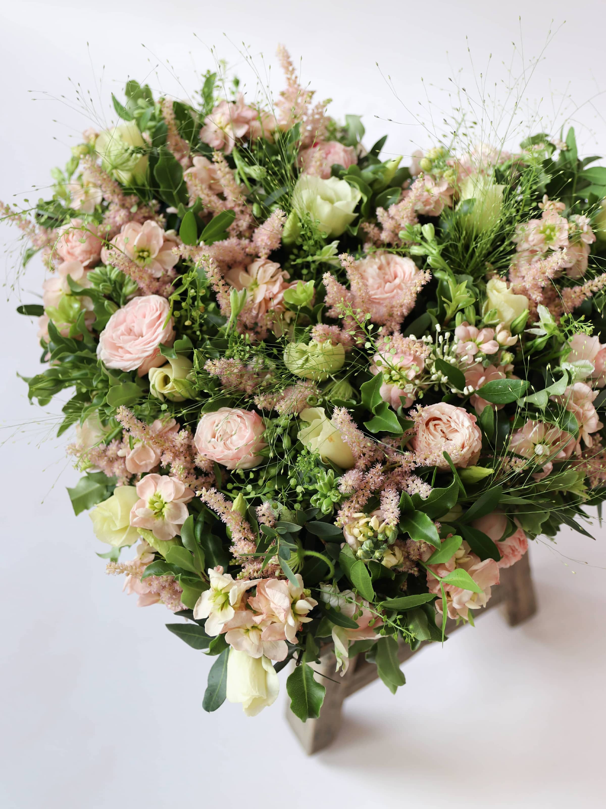 Heart shaped funeral posy with pink and green colours on a white background sitting on a wooden stool