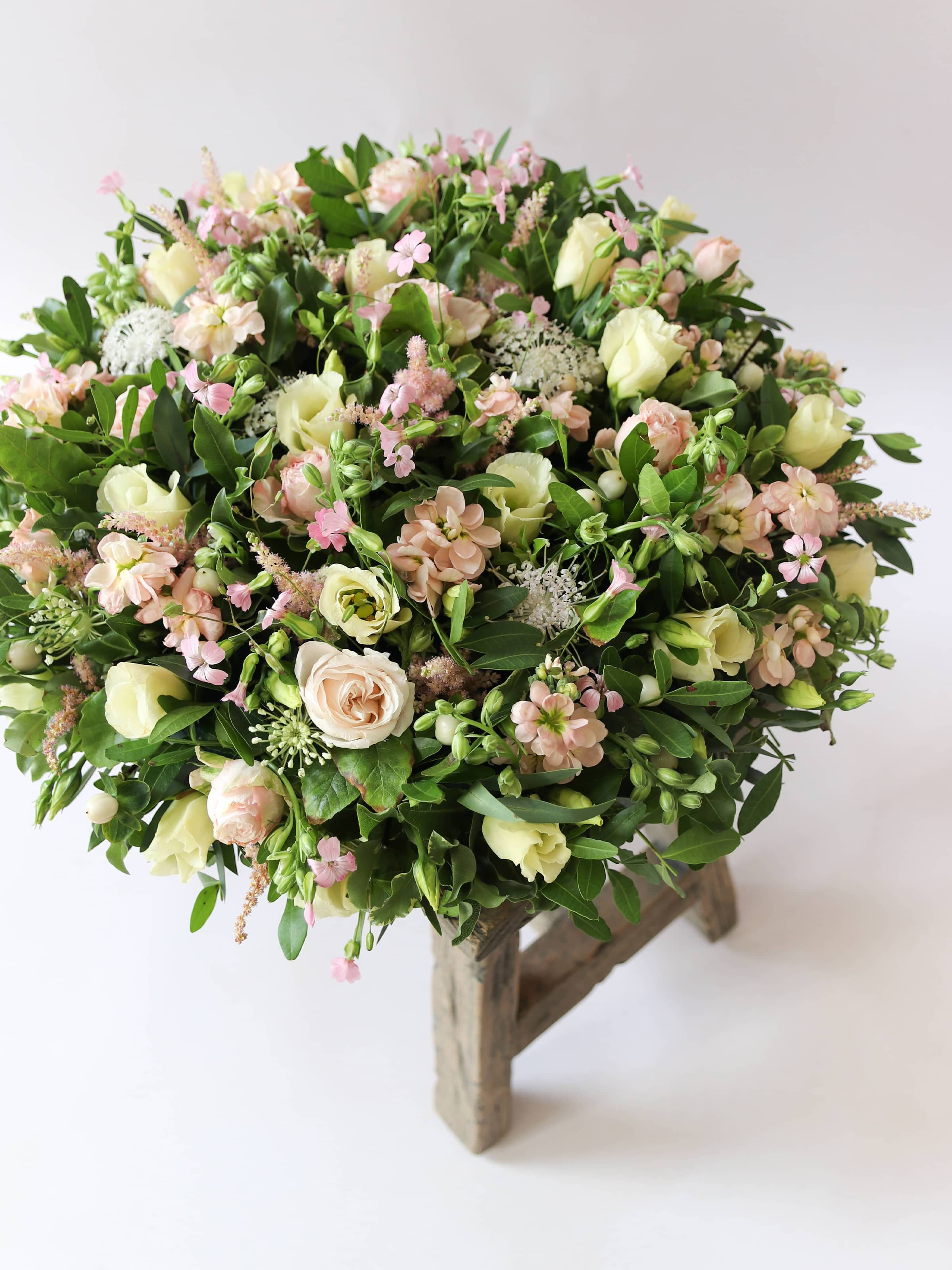 Round funeral posy with white and pastel flowers on a white background sitting on a wooden stool