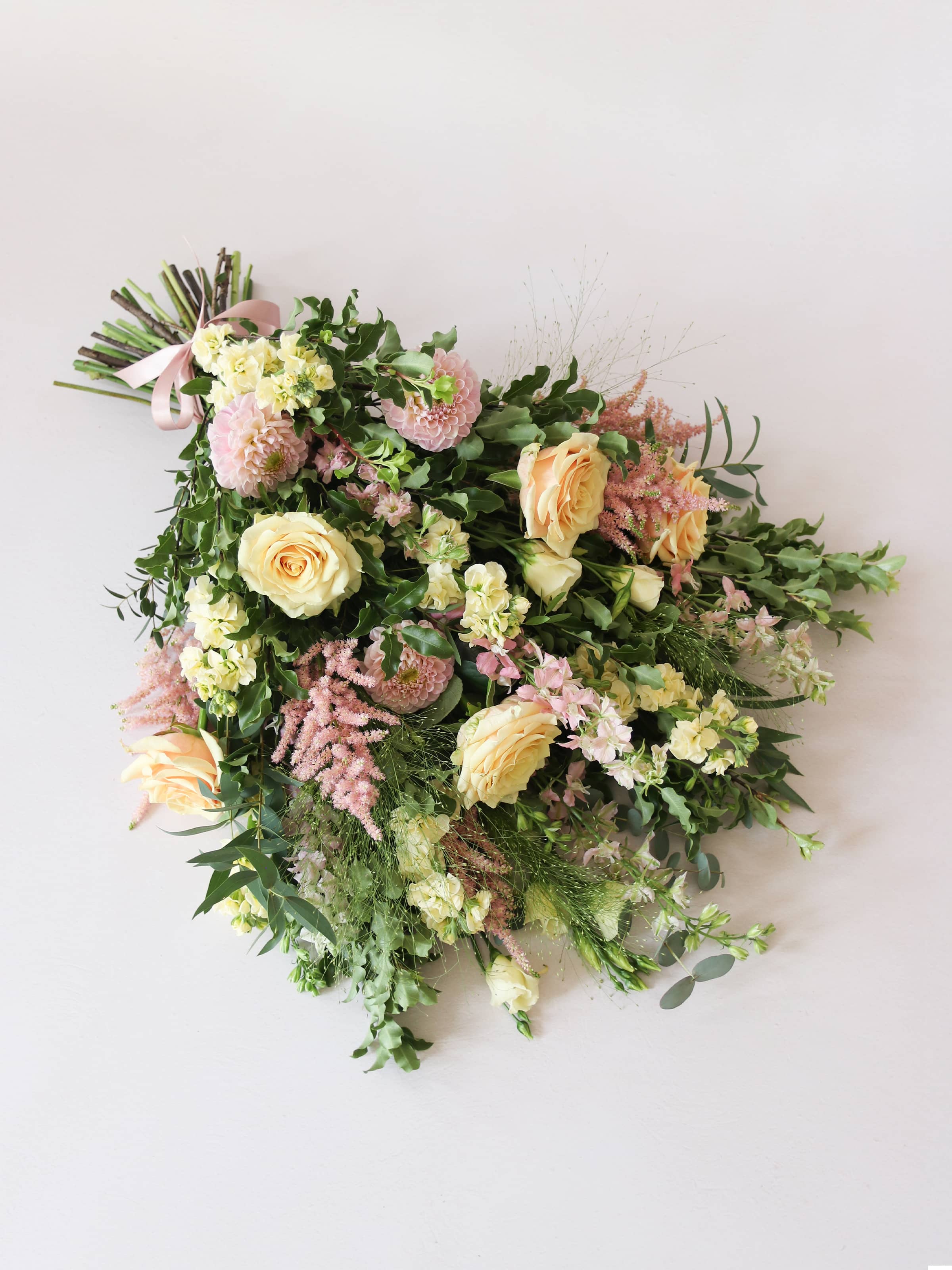 A top down view of a pastel tied funeral sheaf containing pastel pink, peach and yellow flowers, tied with a satin pink ribbon.