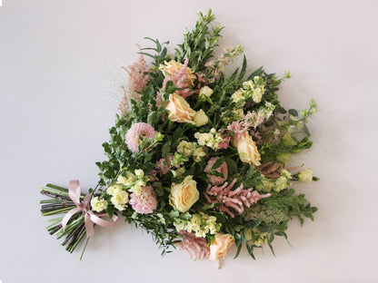 A top down view of a pastel tied funeral sheaf containing pastel pink, peach and yellow flowers, tied with a satin pink ribbon.