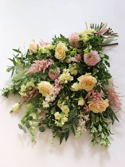 A top down view of a pastel tied funeral sheaf containing pastel pink, peach and yellow flowers, tied with a satin pink ribbon.