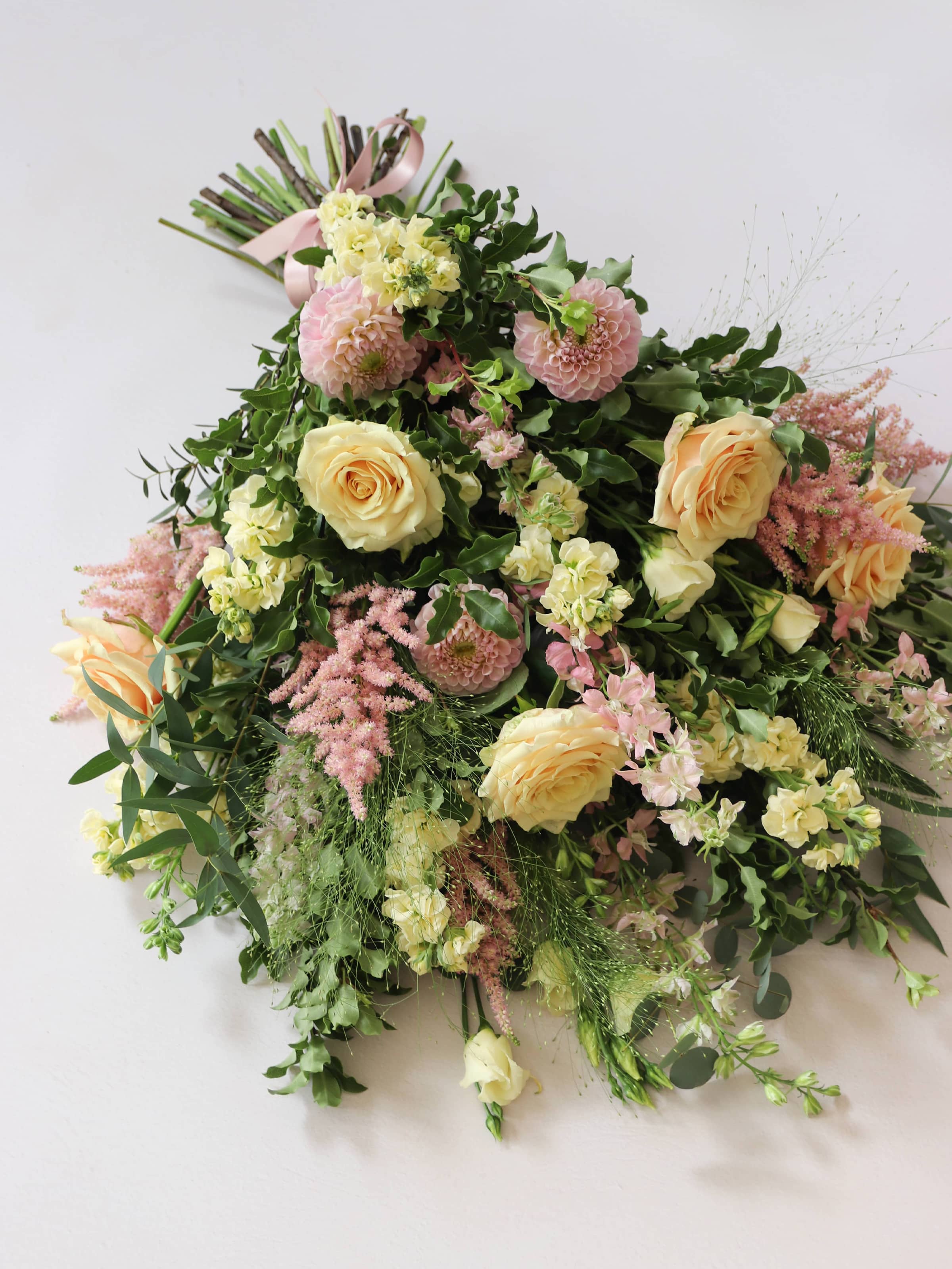A close up image of a pastel tied funeral sheaf containing pastel pink, peach and yellow flowers, tied with a satin pink ribbon.