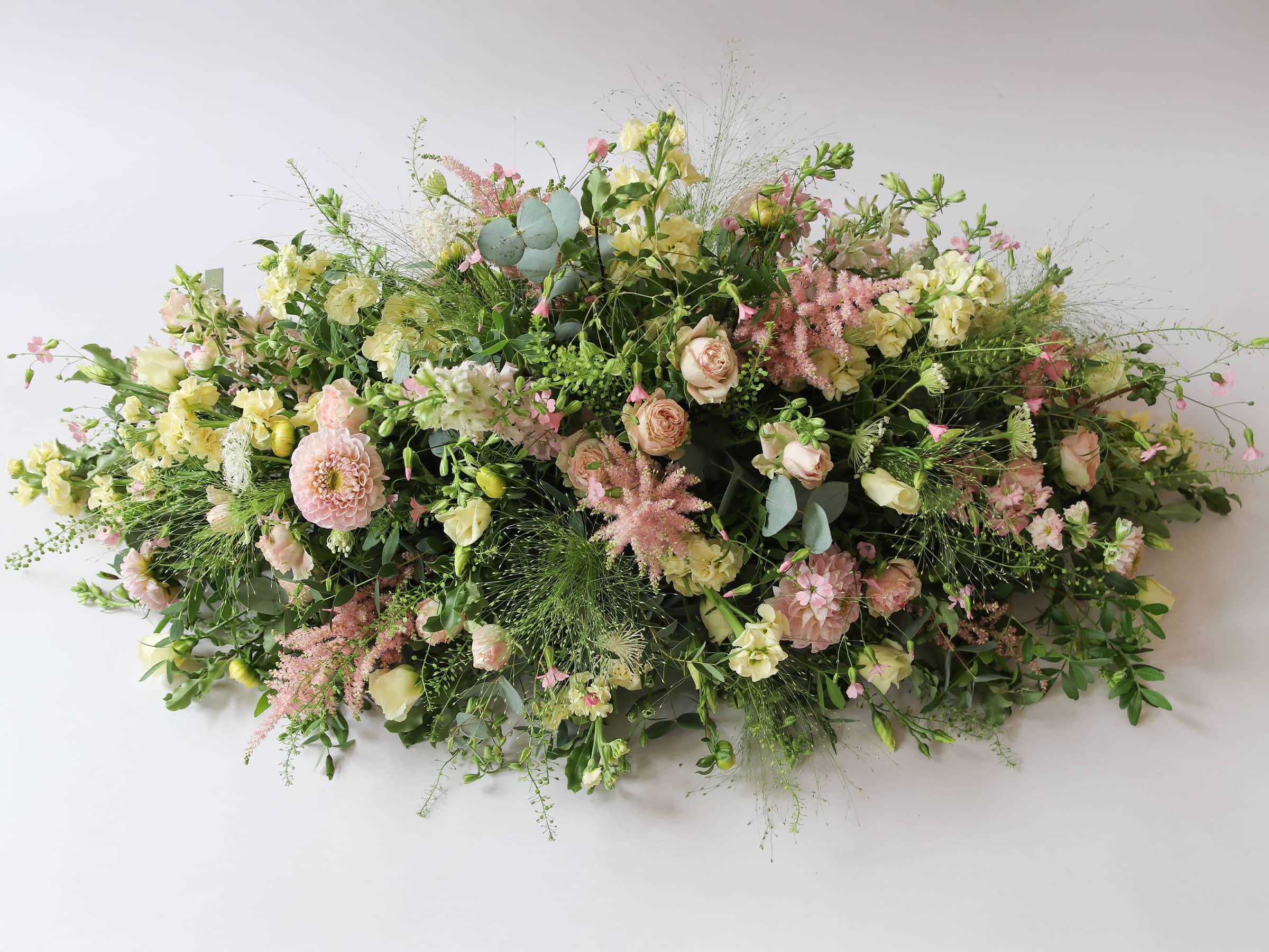 A top view of a 3ft coffin spray featuring pastel pink, yellow and peach flowers and foliage. The funeral arrangement is sitting against a neutral background and features Roses, Astilbe, Stocks and Lisianthus. 
