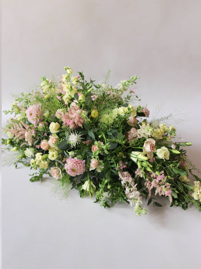 A side view of a 3ft coffin spray featuring pastel pink, yellow and peach flowers and foliage. The funeral arrangement is sitting against a neutral background and features Roses, Astlibe, Stocks and Lisianthus. 