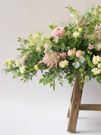A side view of a the pointed end of a 3ft coffin spray featuring pastel pink, yellow and peach flowers and foliage. The funeral arrangement is sitting on a rustic wooden stool against a neutral background and features Roses, Astlibe, Stocks and Lisianthus. 