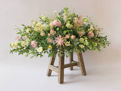 A side view of a 3ft coffin spray featuring pastel pink, yellow and peach flowers and foliage. The funeral arrangement is sitting on a rustic wooden stool against a neutral background and features Roses, Astlibe, Stocks and Lisianthus. 