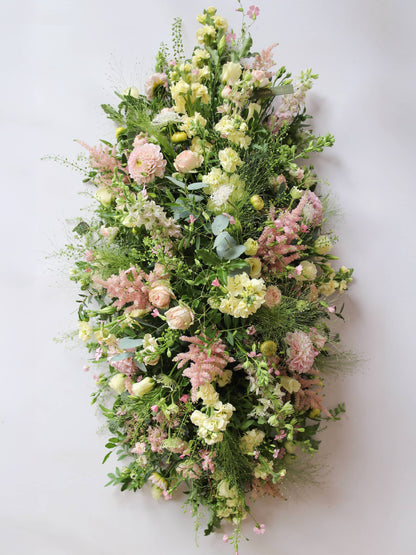 A top view of a 3ft coffin spray featuring pastel pink, yellow and peach flowers and foliage. The funeral arrangement is sitting against a neutral background and features Roses, Astilbe, Stocks and Lisianthus. 