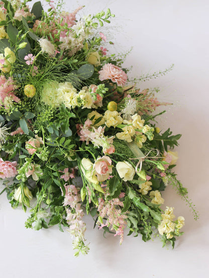 A top view of the pointed end of a 3ft coffin spray featuring pastel pink, yellow and peach flowers and foliage. The funeral arrangement is sitting against a neutral background and features Roses, Astilbe, Stocks and Lisianthus. 