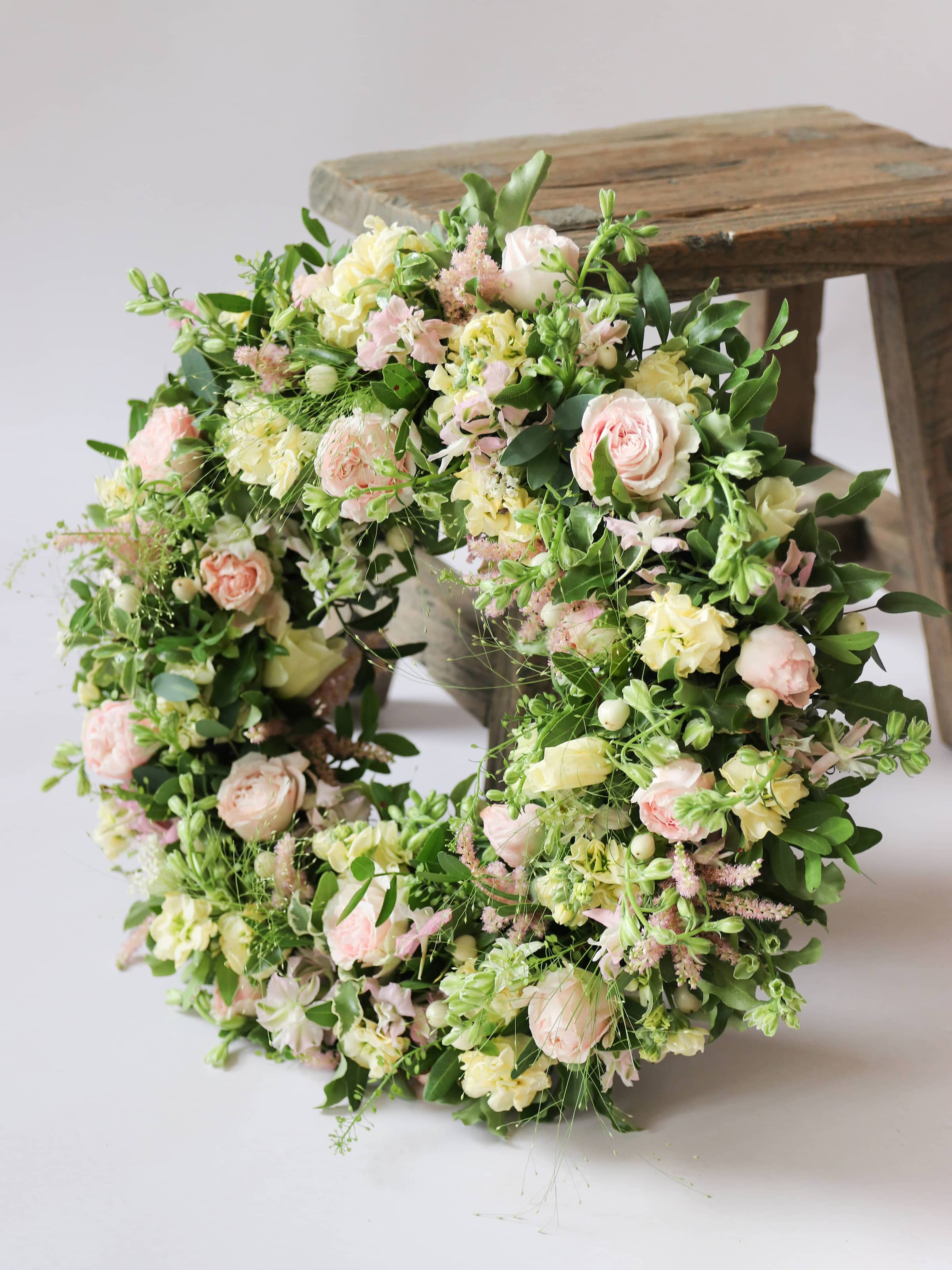 Funeral wreath with pastel and green flowers leaning on a wooden stool against white background