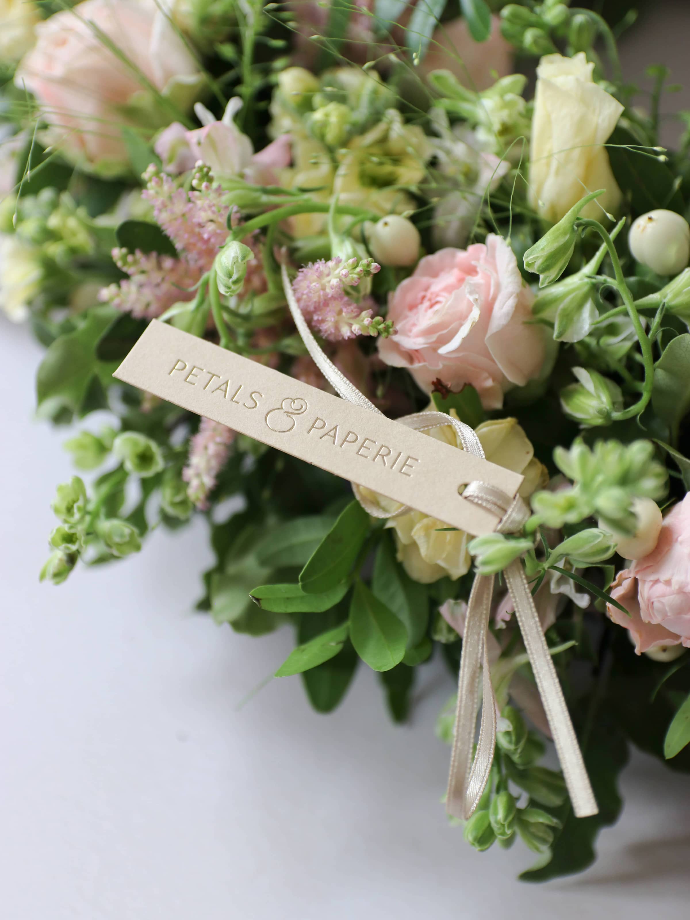 Close up view of a funeral wreath with pastel and green flowers against white background, with a beige and gold fold Petals & Paperie brand tag