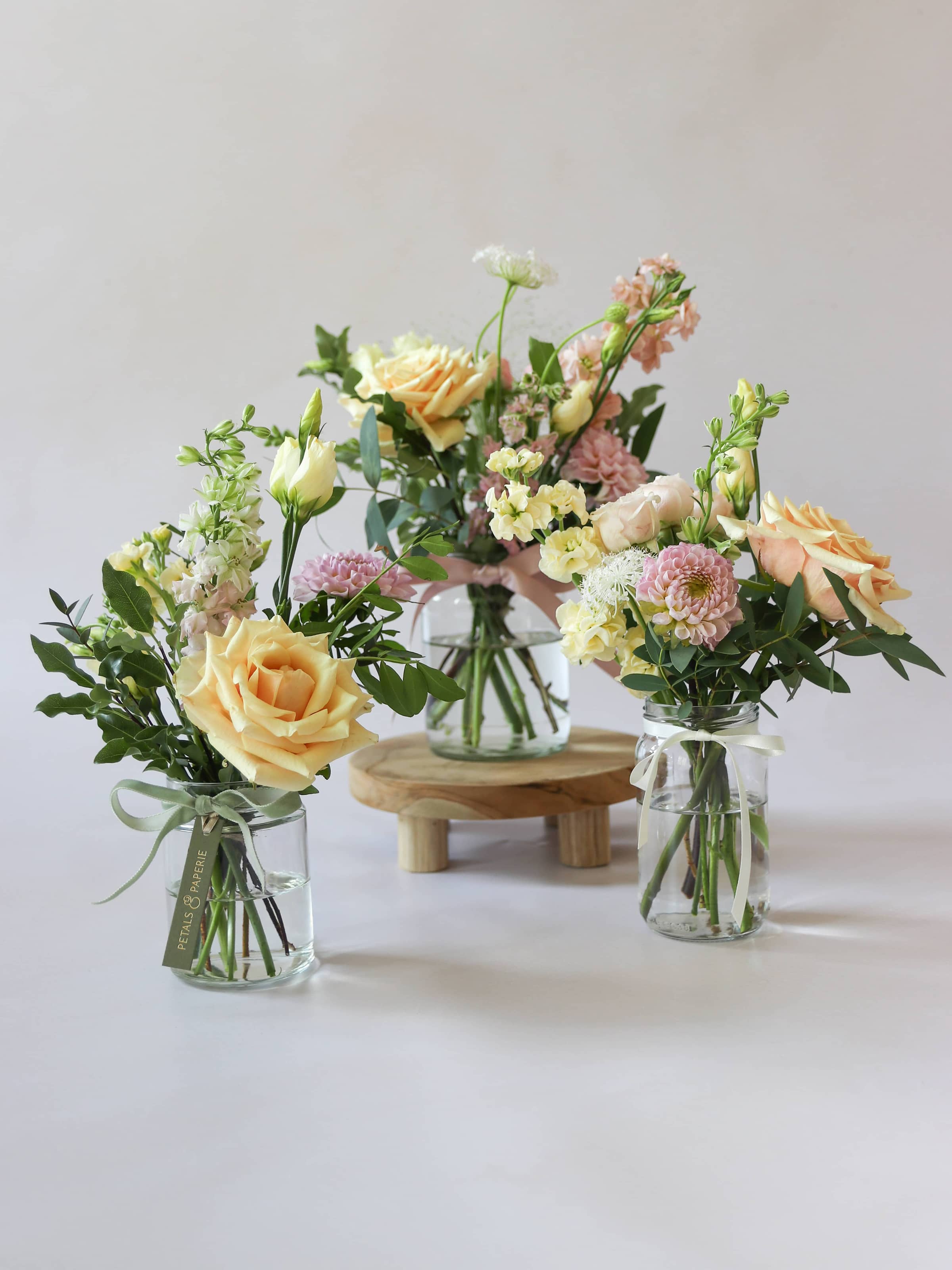 A trio of jam jars with pastel coloured flowers sitting on a wooden stool against a light background