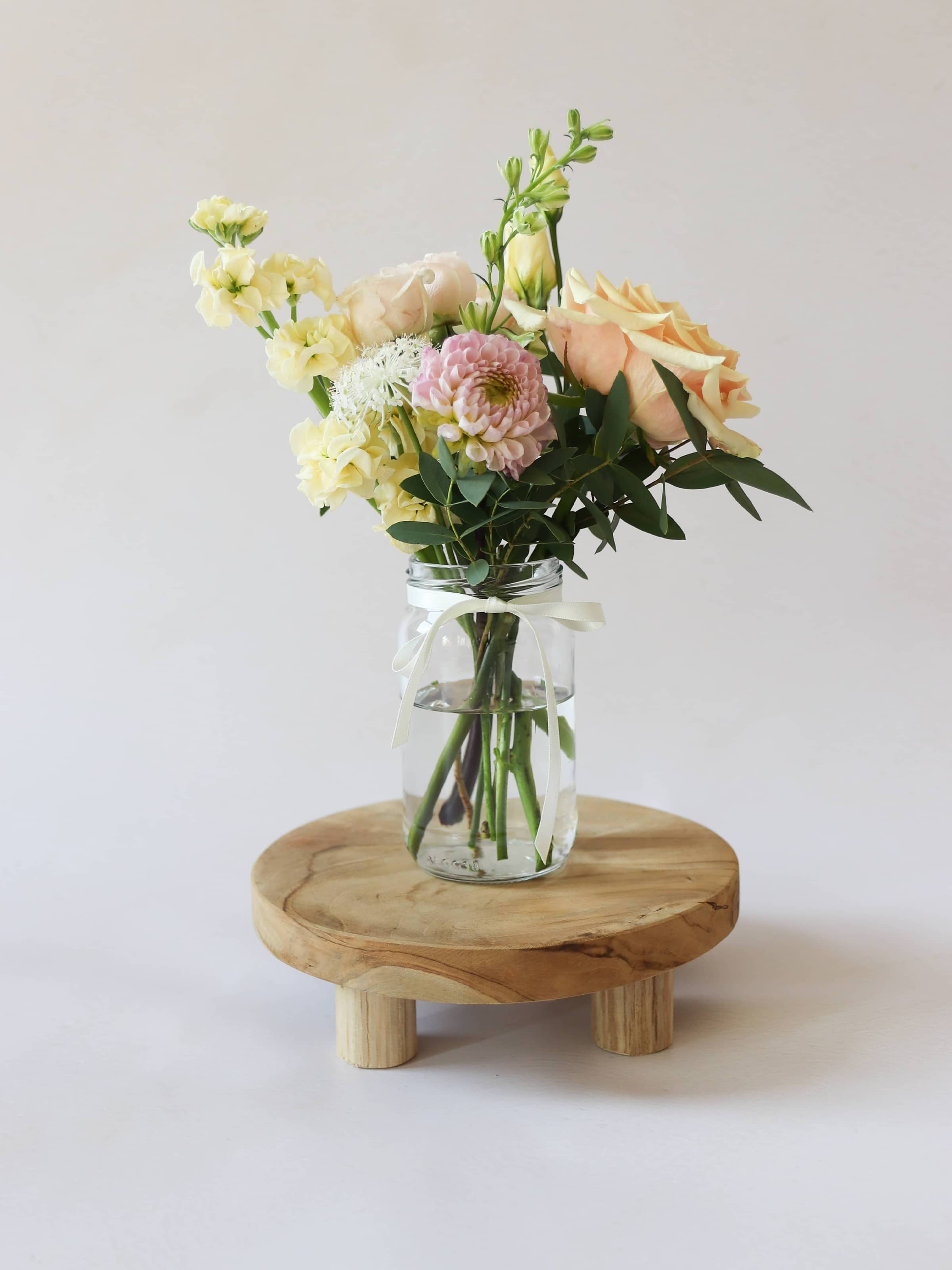 A single jam jar with pastel coloured flowers sitting on a wooden stool against a light background