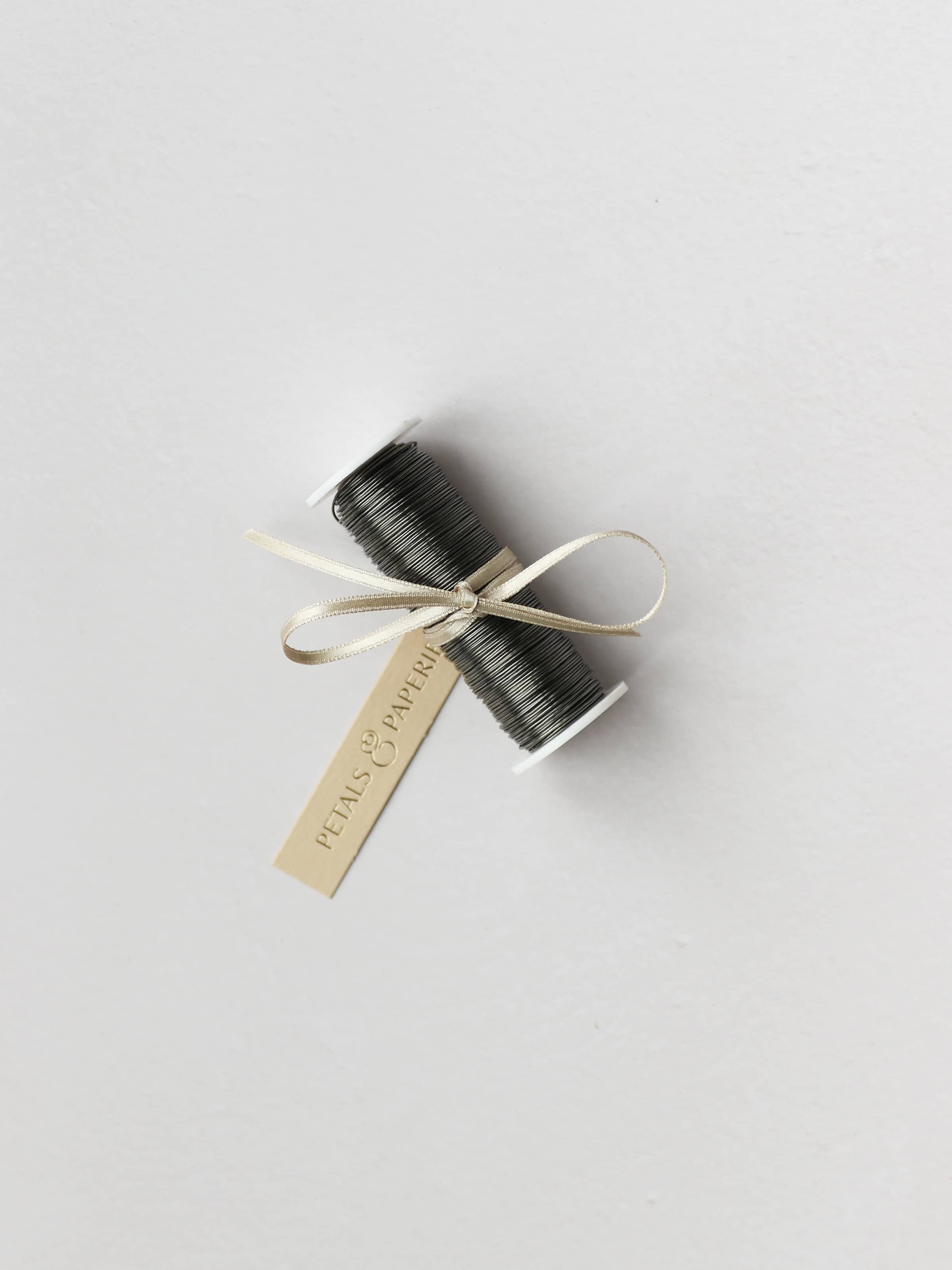 A spool of silver reel wire for Christmas wreath making, tied with a cream satin ribbon and Petals & Paperie gift tag on a neutral background.