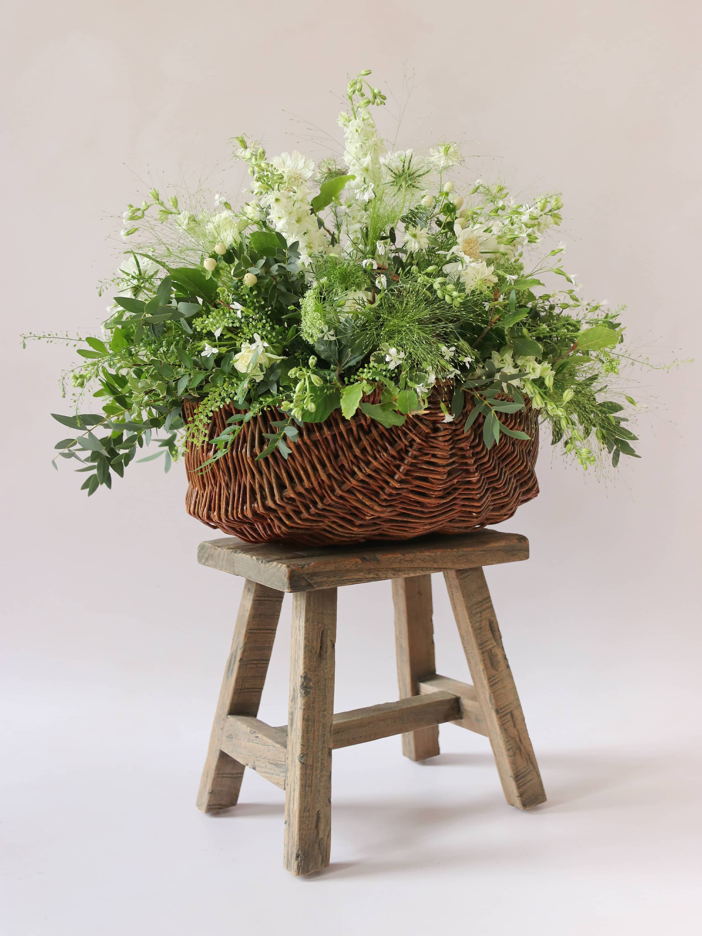A wicker basket arrangement featuring white and green flowers and foliage. The basket arrangement is sitting on a rustic wooden stool against a neutral background and features white Roses, Ammi, Stocks and Lisianthus. 