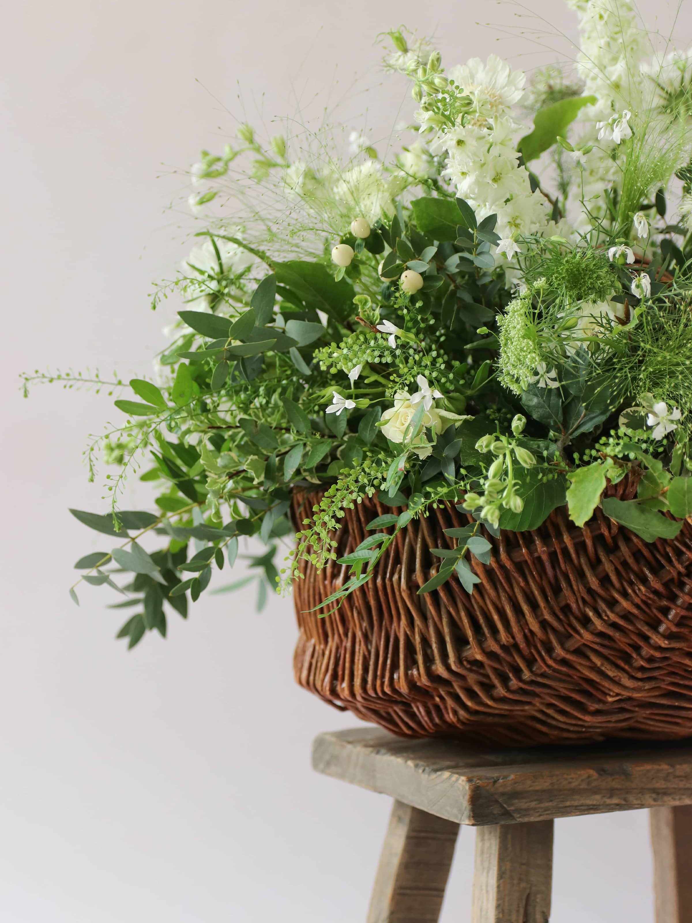 A side view of a wicker basket arrangement featuring white and green flowers and foliage. The basket arrangement is sitting on a rustic wooden stool against a neutral background and features white Roses, Ammi, Stocks and Lisianthus. 