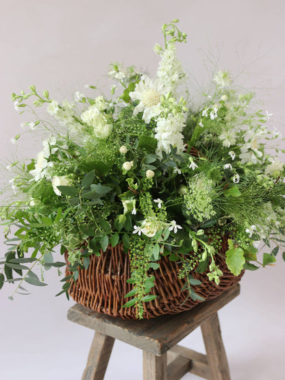 A side view of a wicker basket arrangement featuring white and green flowers and foliage. The basket arrangement is sitting on a rustic wooden stool against a neutral background and features white Roses, Ammi, Stocks and Lisianthus. 