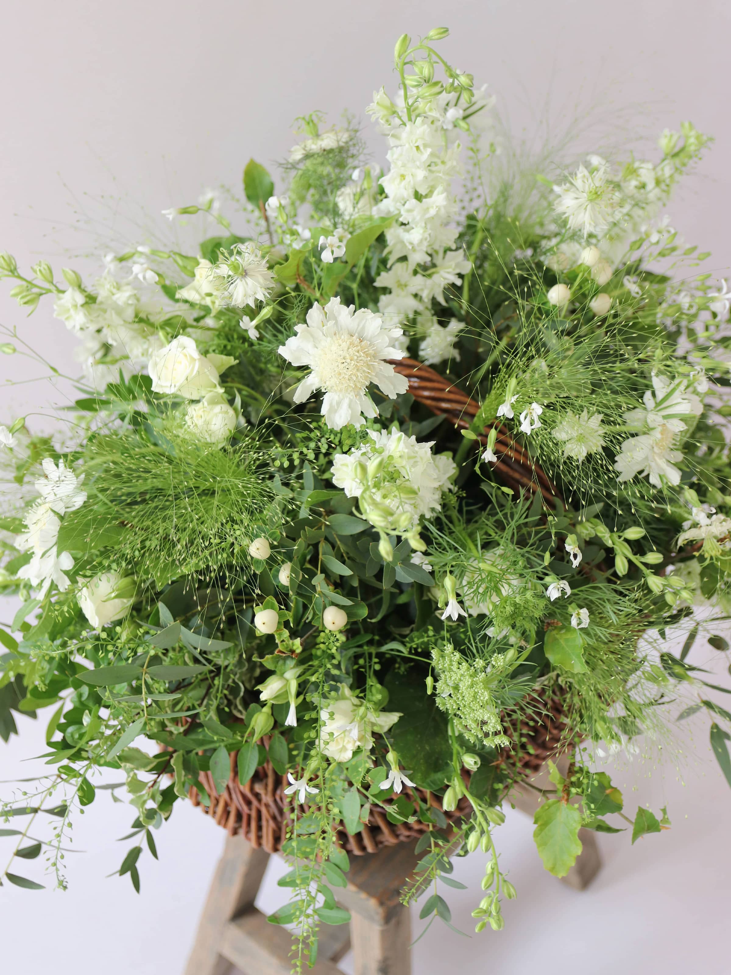 A top view of a wicker basket arrangement featuring white and green flowers and foliage. The basket arrangement is sitting on a rustic wooden stool against a neutral background and features white Roses, Ammi, Stocks and Lisianthus. 