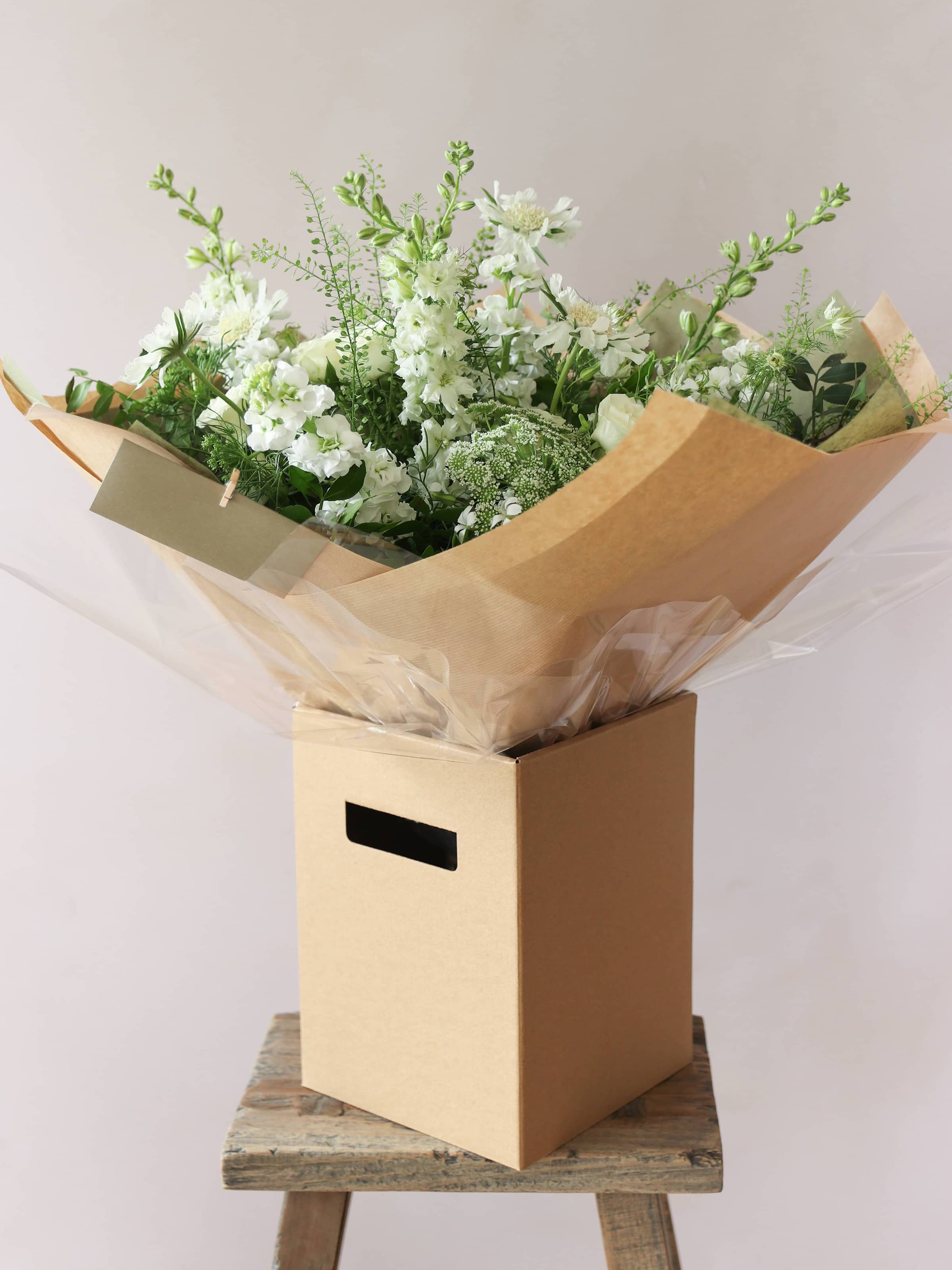 A gift-wrapped bouquet of green and white flowers standing in a kraft brown gift  box. The gift box is sitting on a small wooden stool against a neutral background.