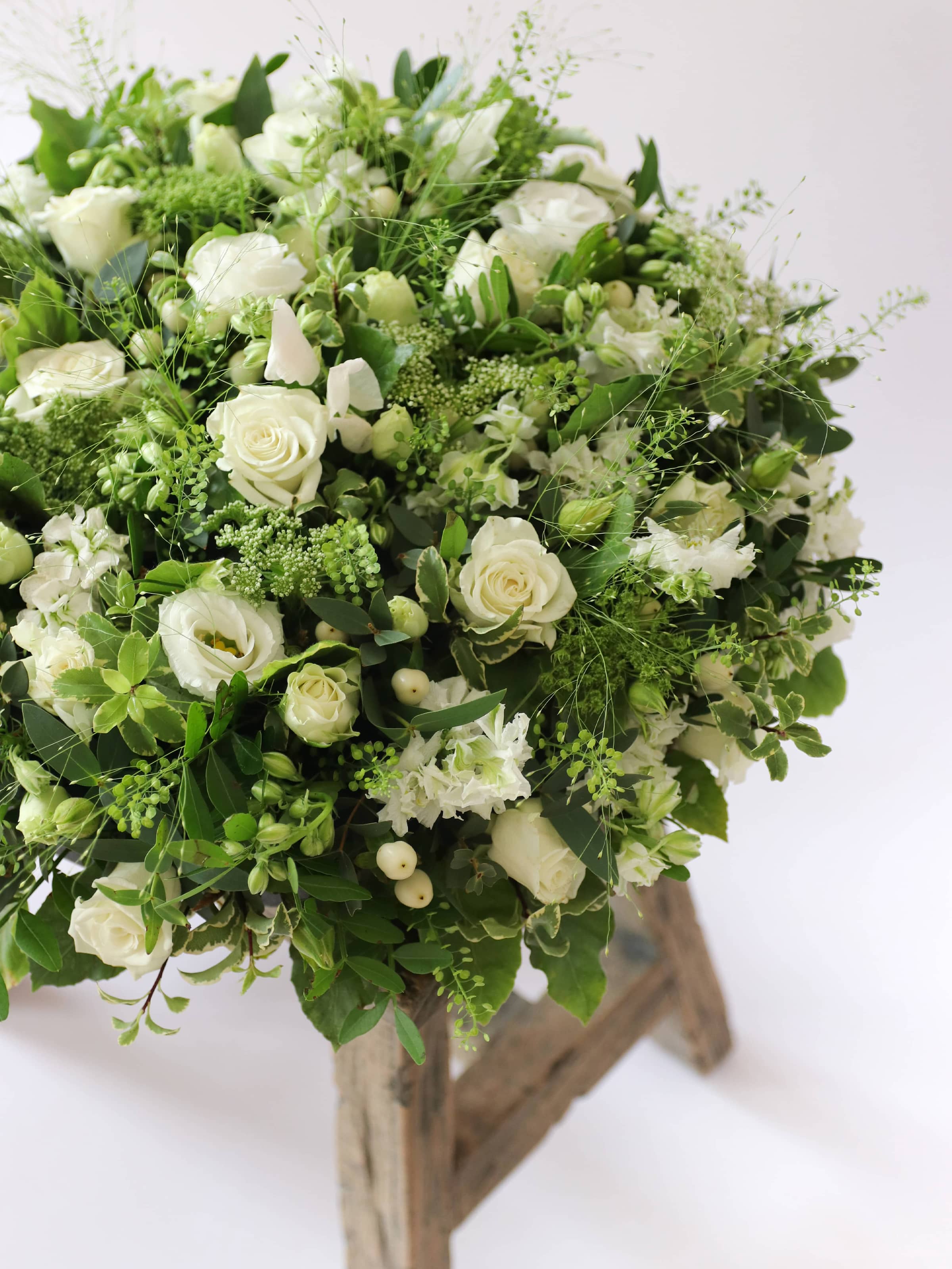 Close up of a round funeral posy with white and green flowers on a white background sitting on a wooden stool