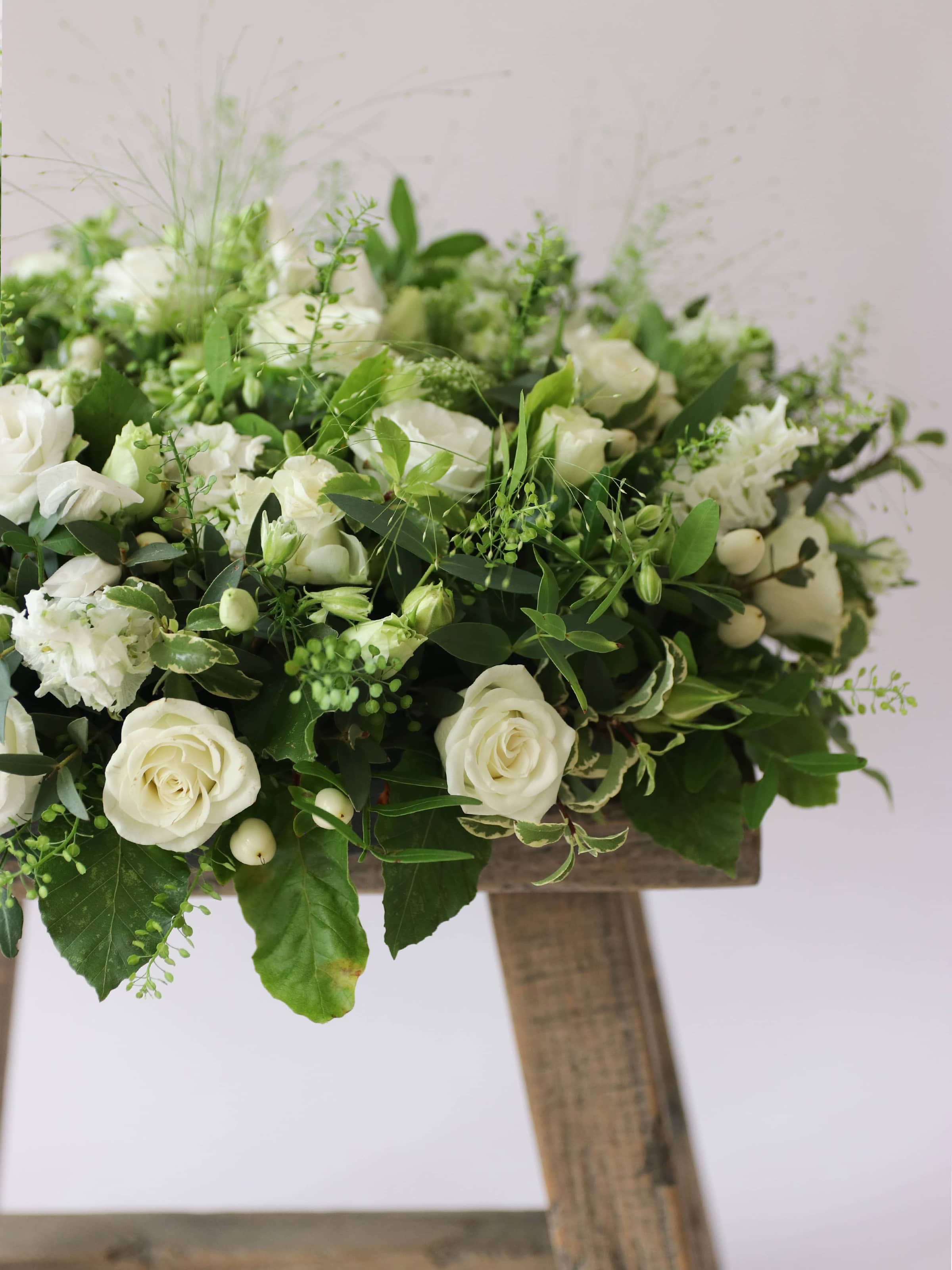 Round funeral posy with white and green flowers on a white background sitting on a wooden stool