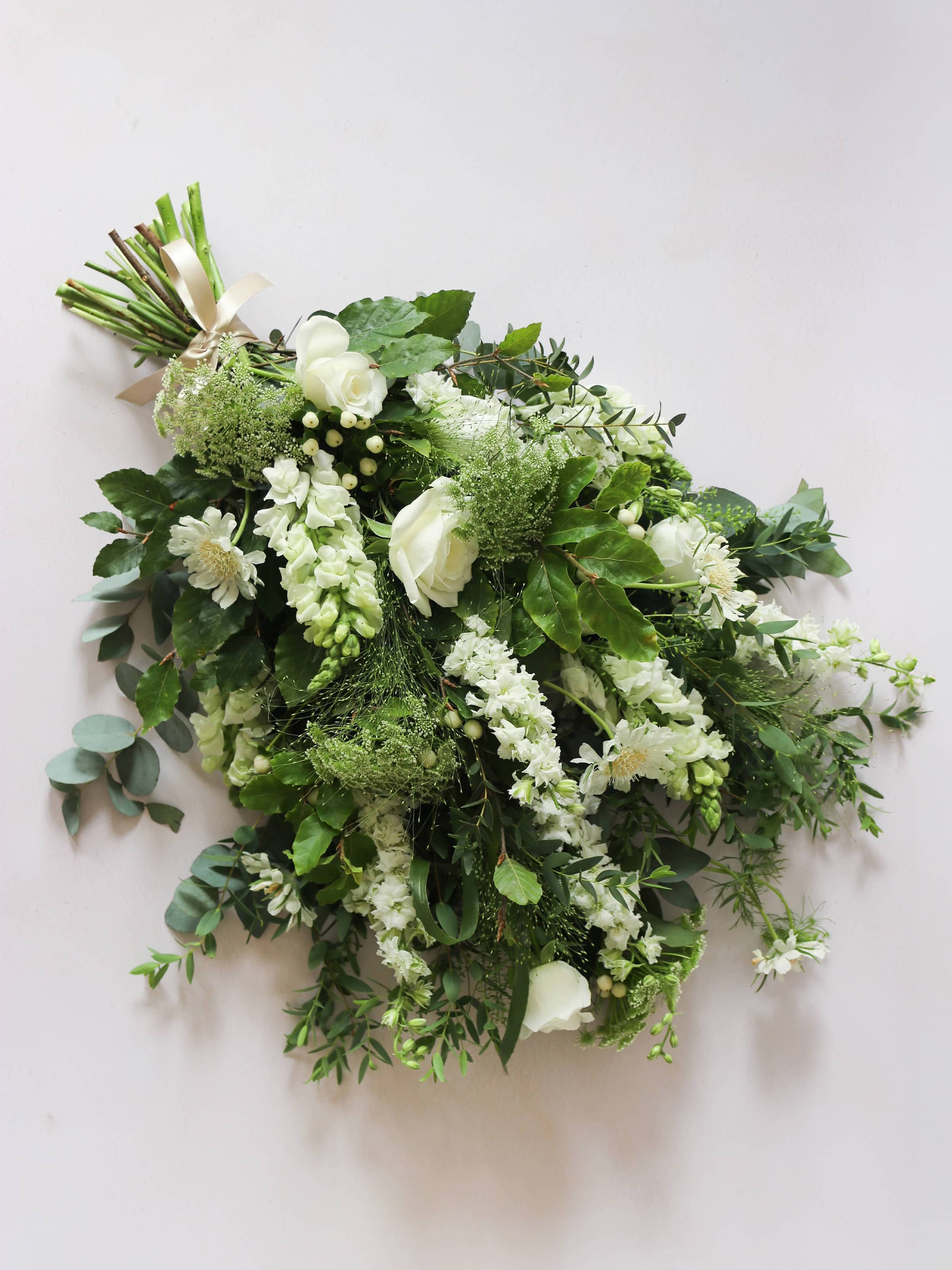 A top down view of a white and green tied funeral sheaf containing white flowers and green foliages, tied with a champagne colour satin ribbon. The sheaf contains Delphiniums, Snapdragons, Ammi and Roses.