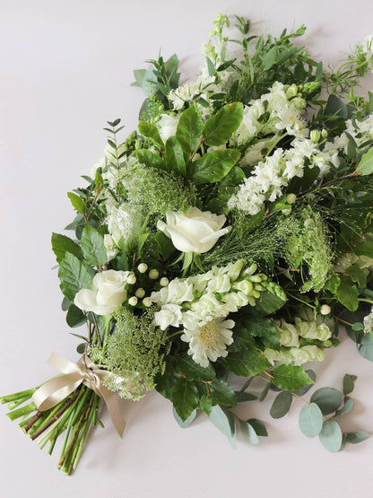 A top down view of a white and green tied funeral sheaf containing white flowers and green foliages, tied with a champagne colour satin ribbon. The sheaf contains Delphiniums, Snapdragons, Ammi and Roses.