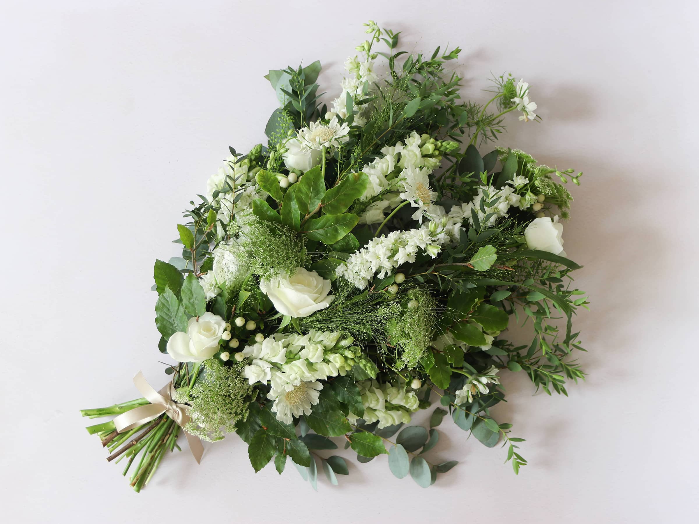 A top down view of a white and green tied funeral sheaf containing white flowers and green foliages, tied with a champagne colour satin ribbon. The sheaf contains Delphiniums, Snapdragons, Ammi and Roses.
