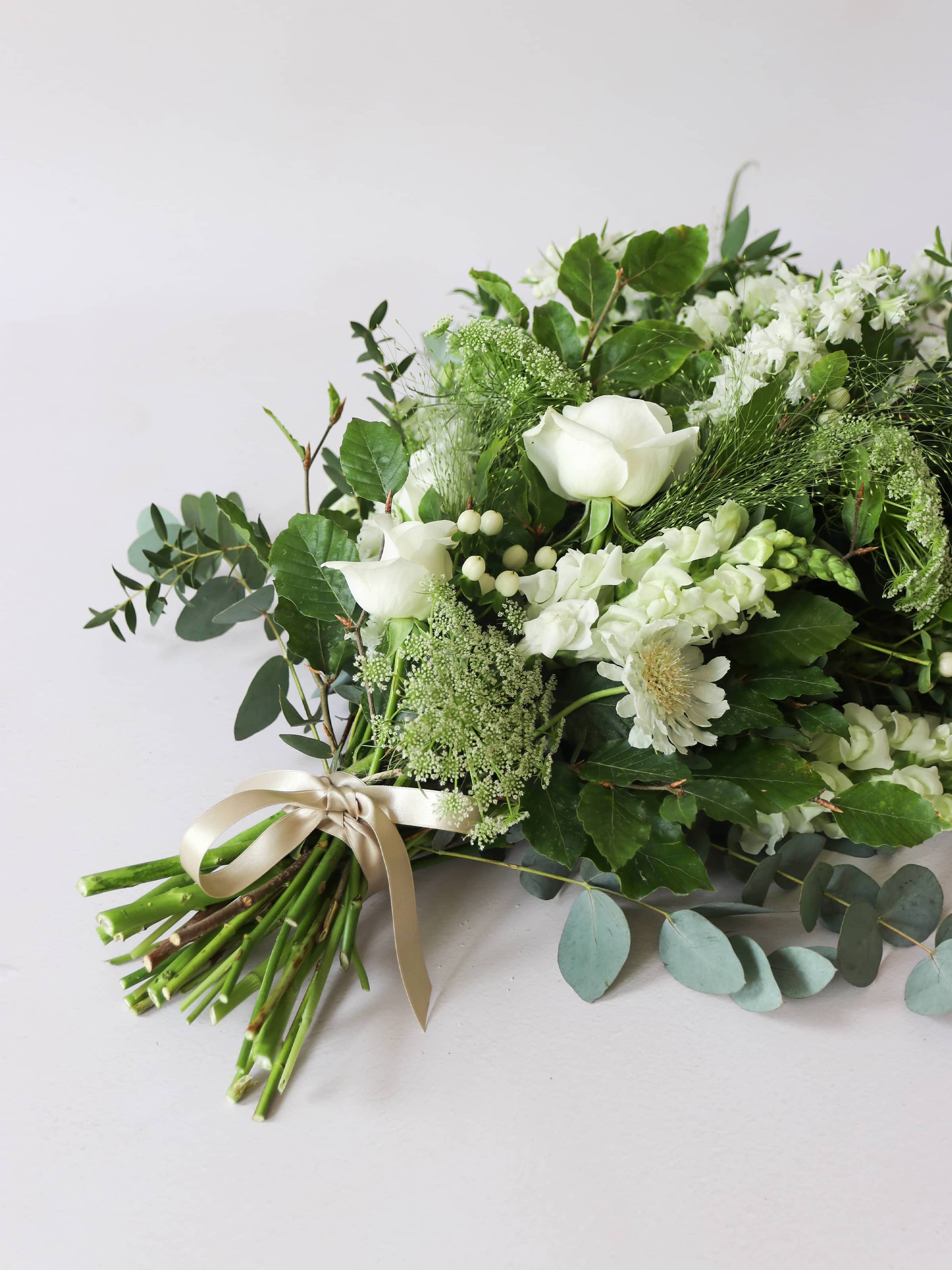 A top down view of a white and green tied funeral sheaf containing white flowers and green foliages, tied with a champagne colour satin ribbon. The sheaf contains Delphiniums, Snapdragons, Ammi and Roses.