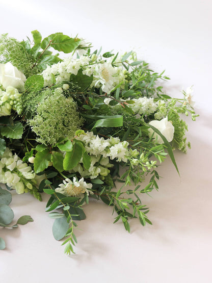 A detail view of a white and green tied funeral sheaf containing white flowers and green foliages, tied with a champagne colour satin ribbon. The sheaf contains Delphiniums, Snapdragons, Ammi and Roses.