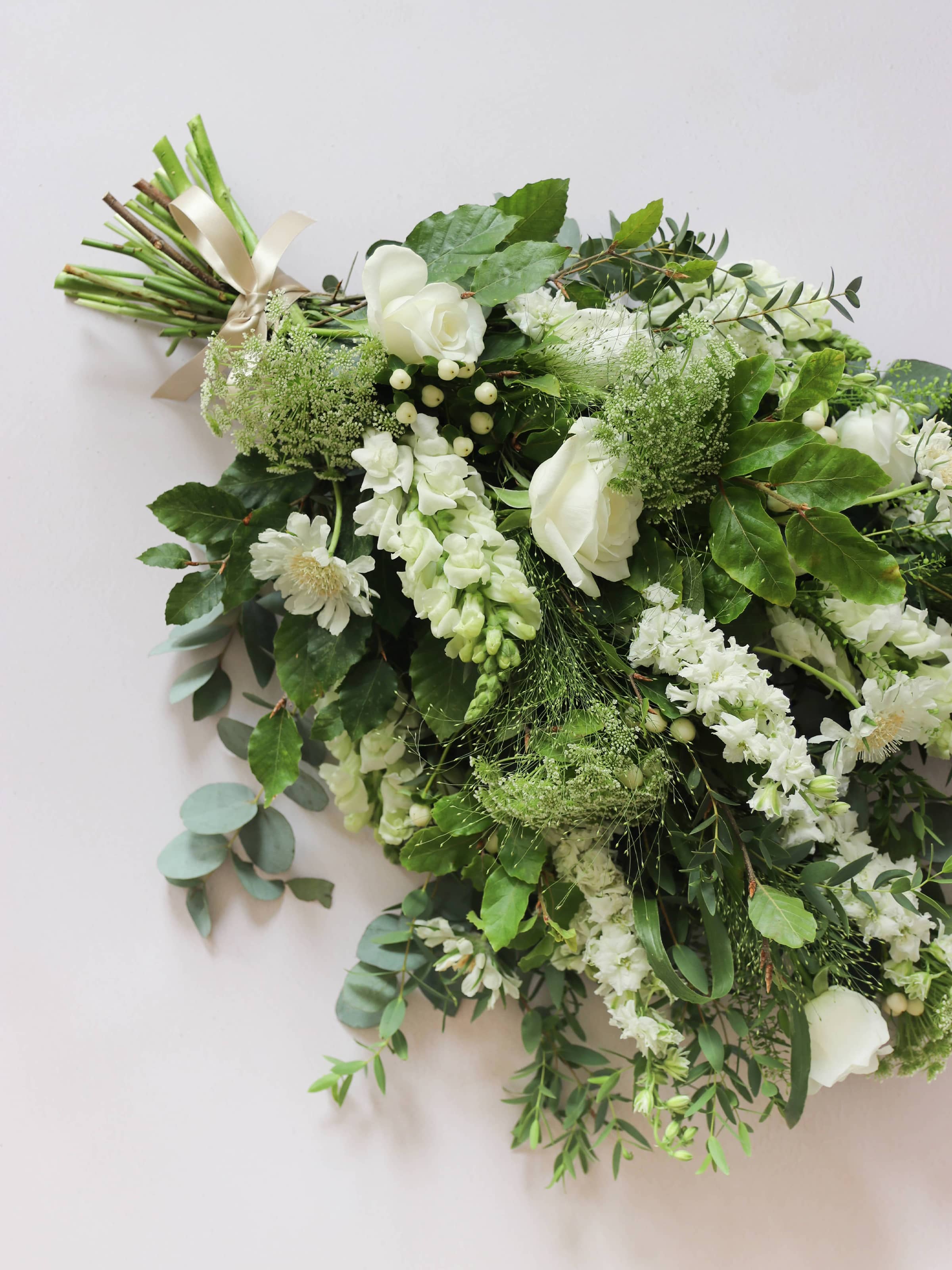 A top down view of a white and green tied funeral sheaf containing white flowers and green foliages, tied with a champagne colour satin ribbon. The sheaf contains Delphiniums, Snapdragons, Ammi and Roses.