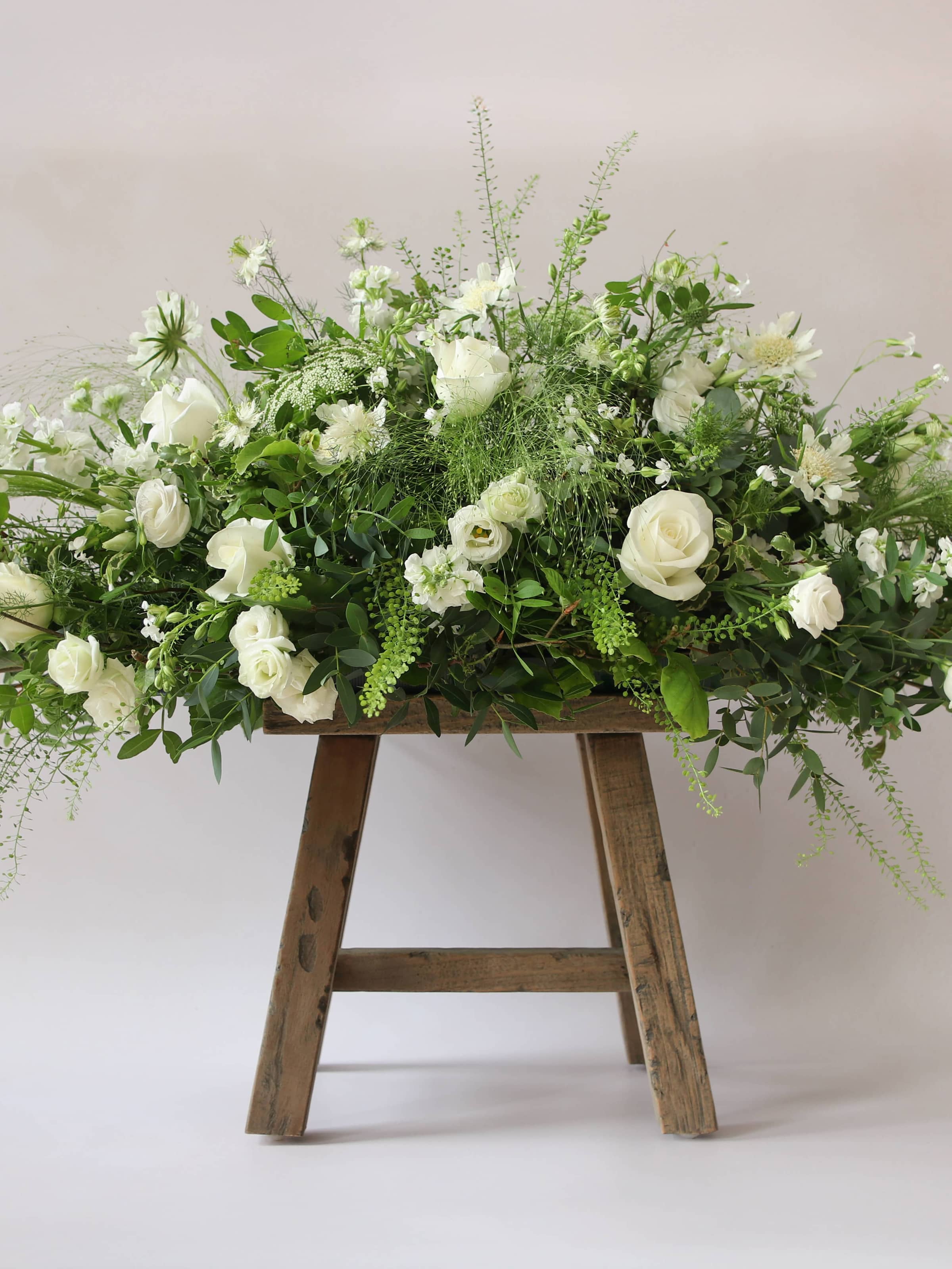 A close up of the middle section of a 3ft coffin spray featuring white and green flowers and foliage. The funeral arrangement is sitting on a rustic wooden stool against a neutral background and features white Roses, Ammi, Stocks and Lisianthus. 