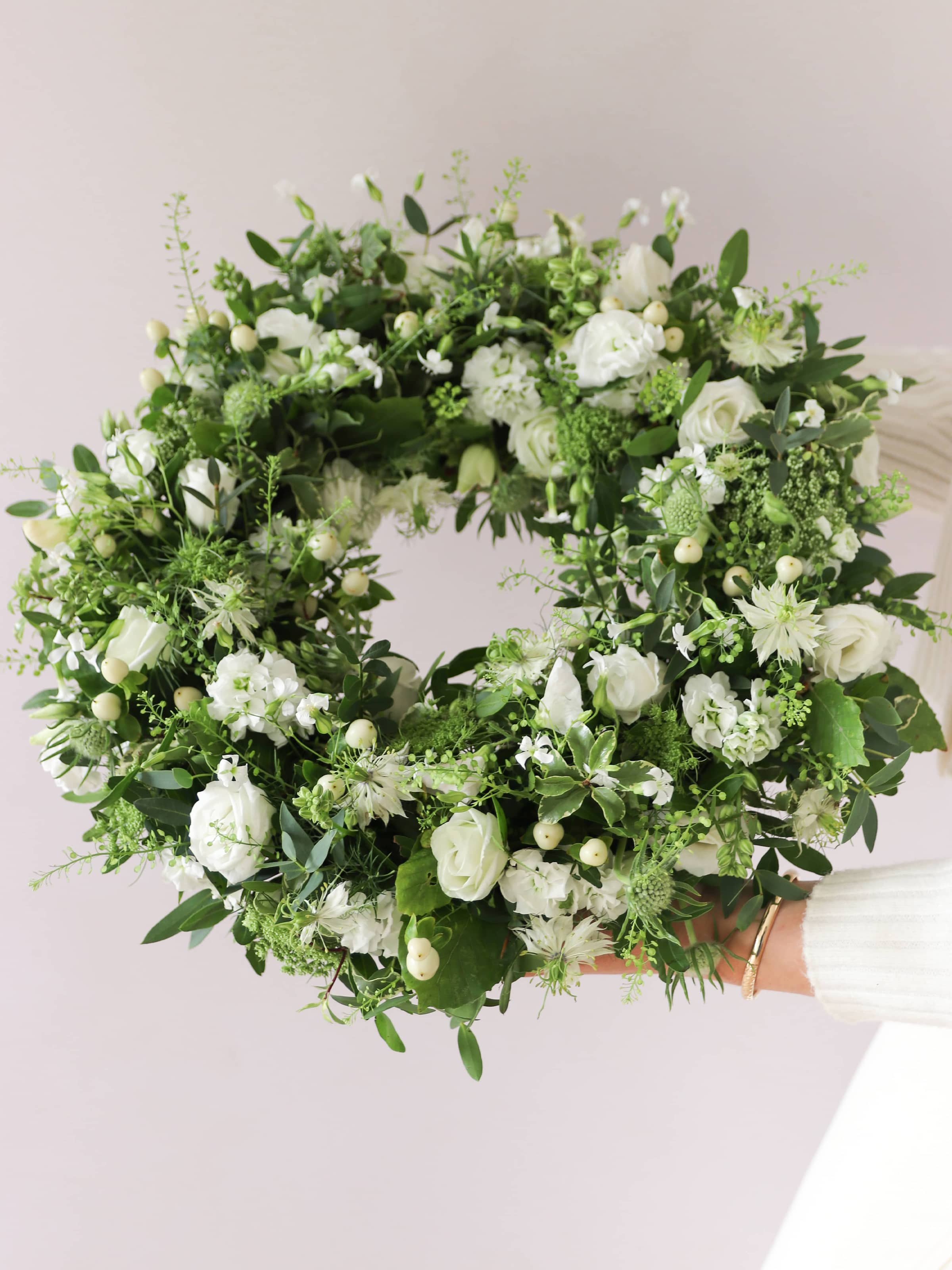 Funeral wreath with white and green flowers being held by a women in neutral clothing against a white background