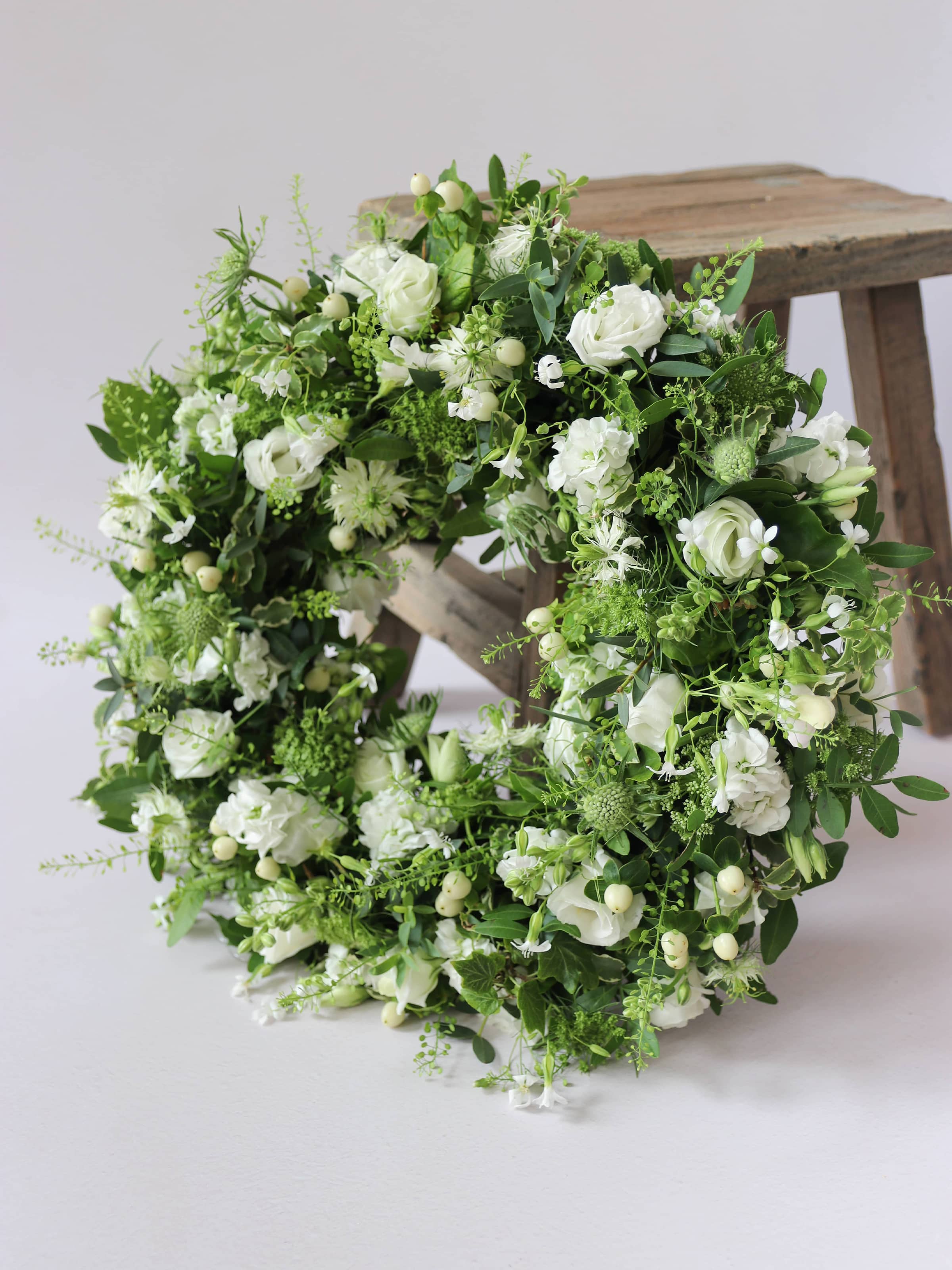 Funeral wreath with white and green flowers leaning against a wooden stool against a white background