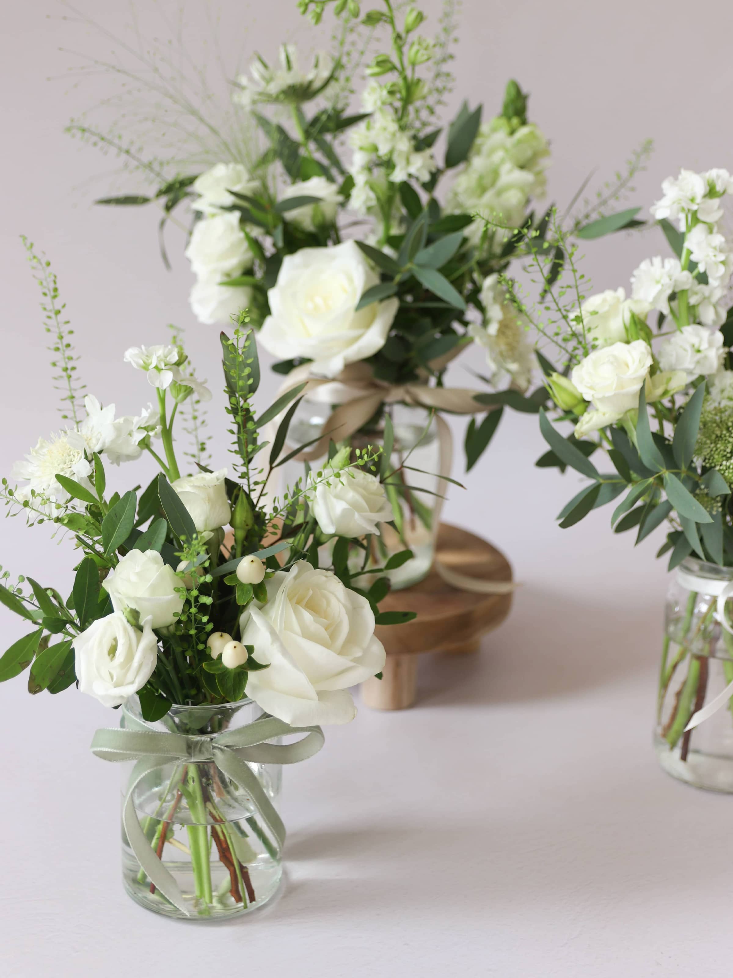 Close up of three glass jars filled with white flowers and greenery on a light background