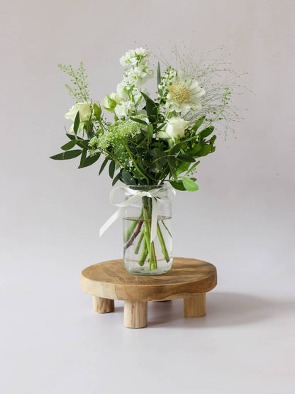 A single jam jar with white and green flowers sitting on a wooden stool against a light background