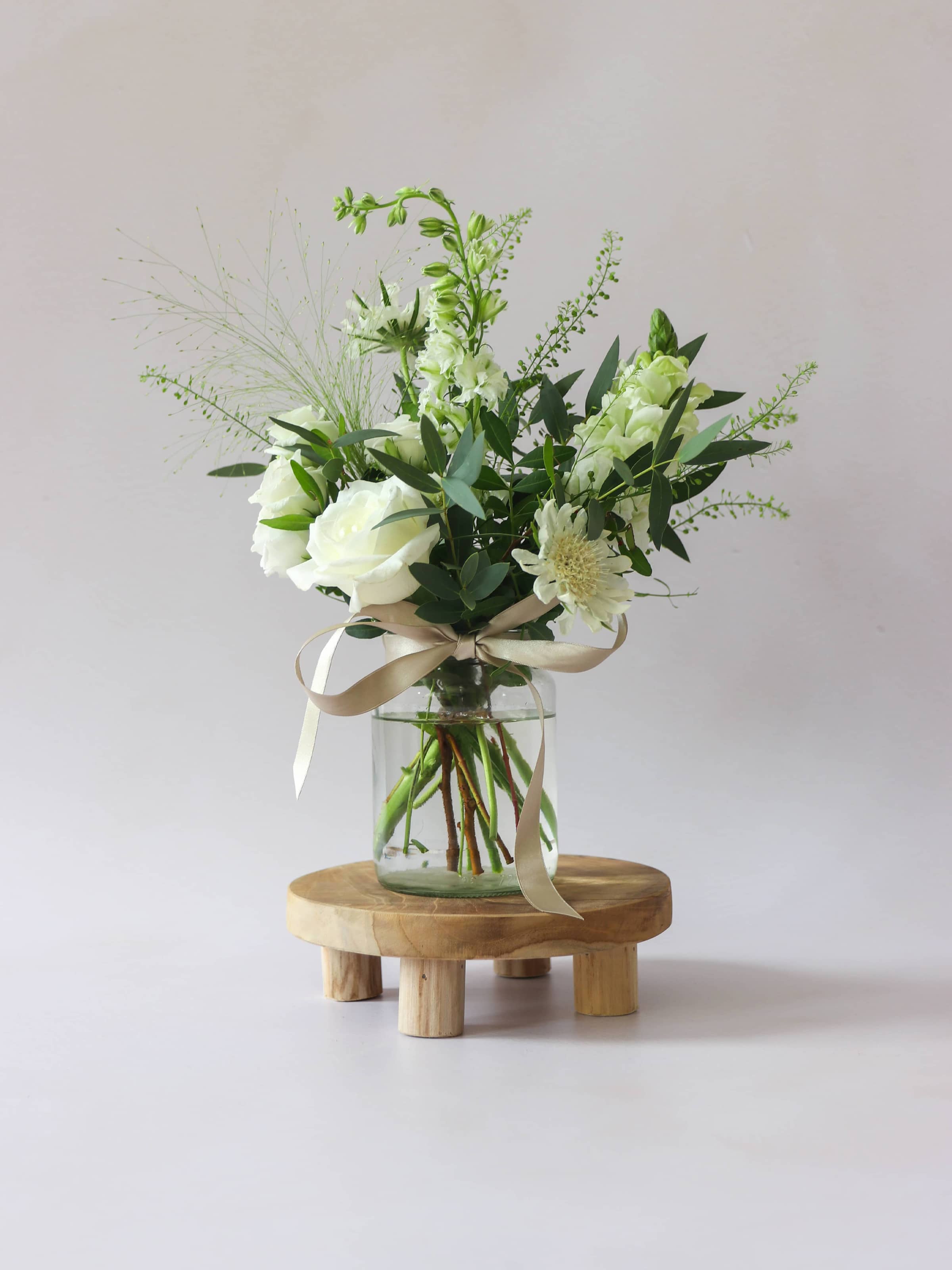 A single jam jar with white and green flowers sitting on a wooden stool against a light background