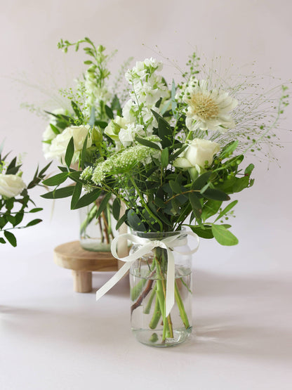 Close up of a single jam jar with white and green flowers sitting on a wooden stool against a light background