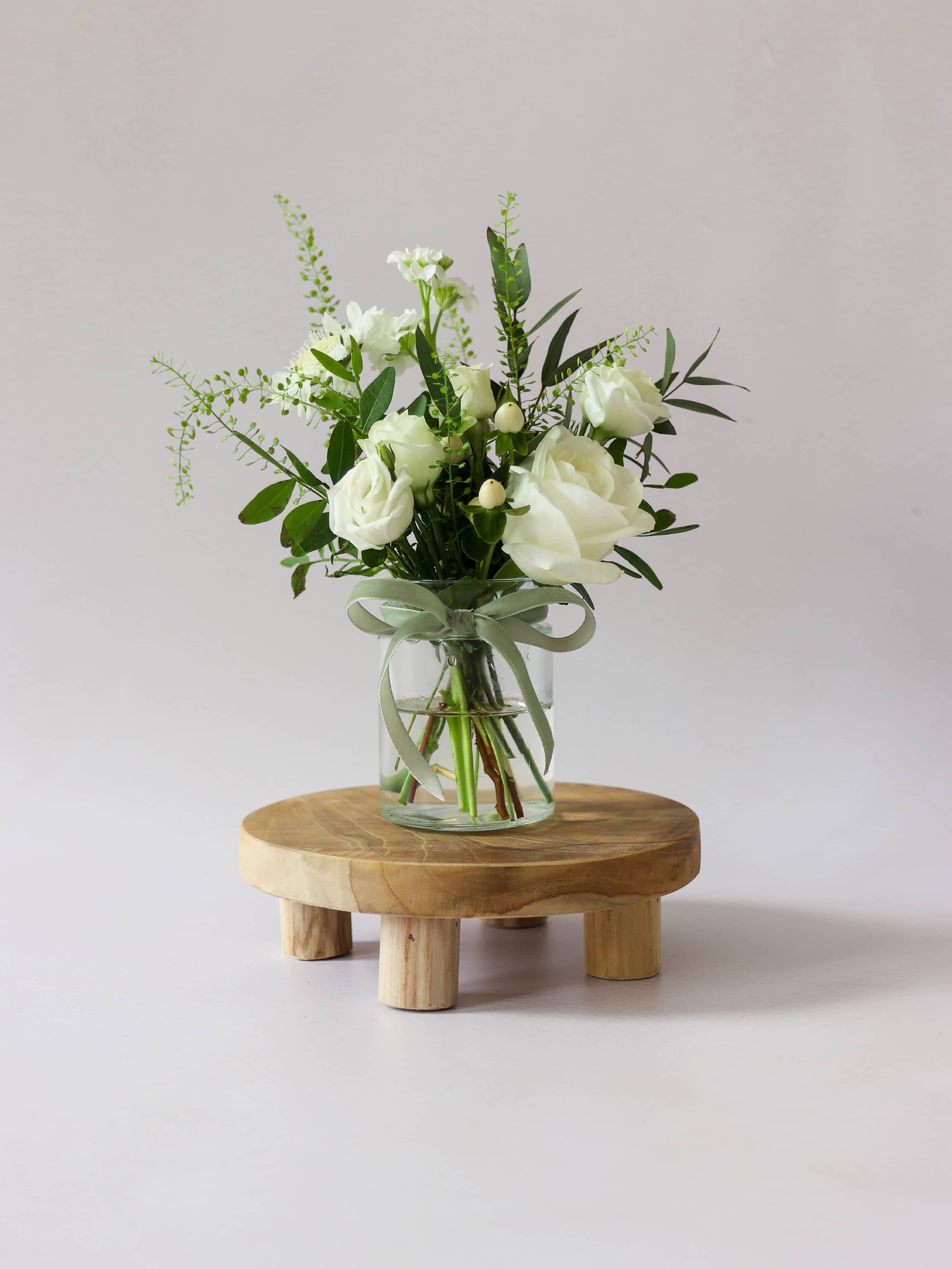 A single jam jar with white and green flowers sitting on a wooden stool against a light background